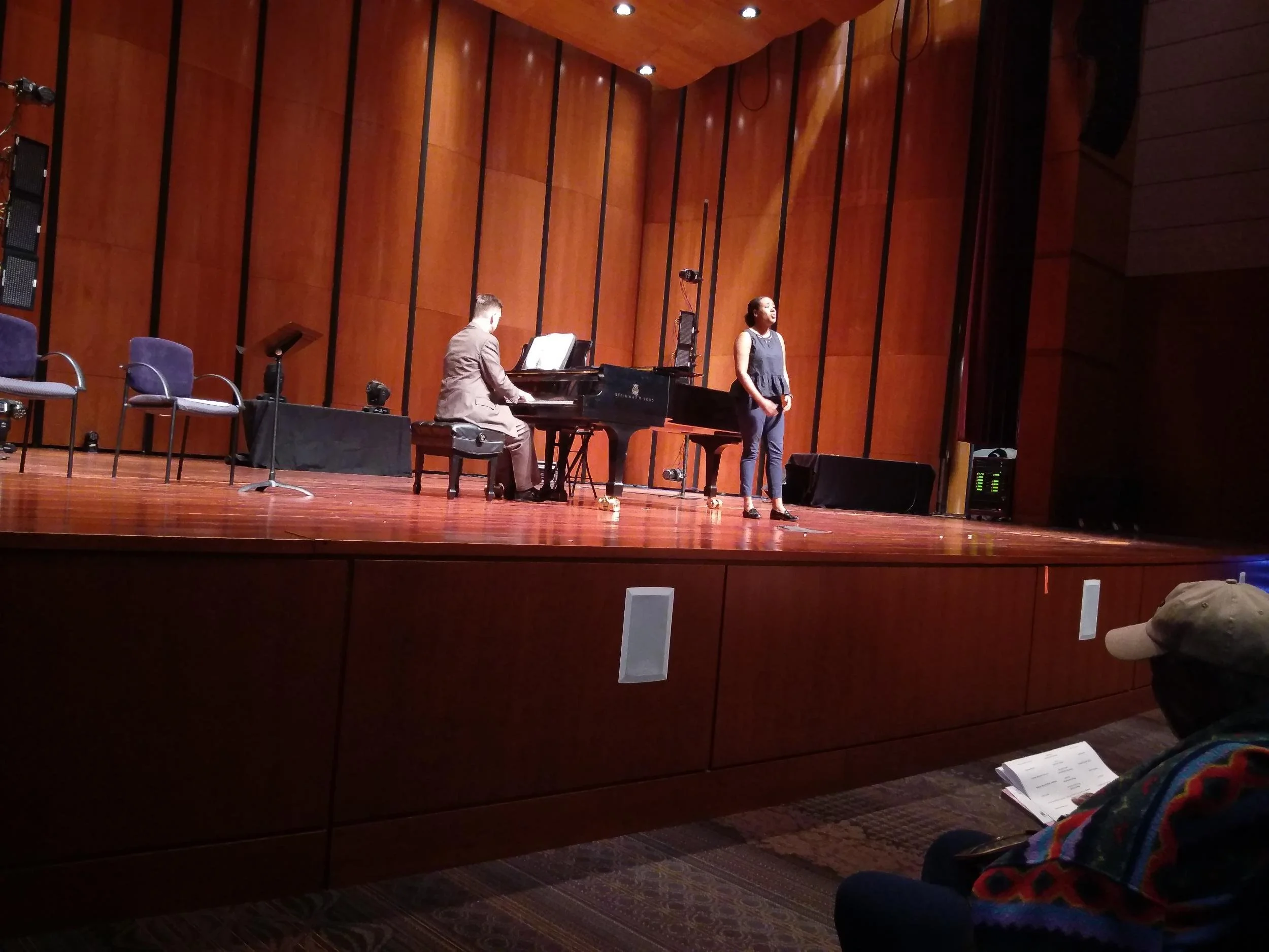 A person singing on stage with a pianist accompanying, in a concert hall with wooden walls. There are empty chairs and a microphone stand on the stage.
