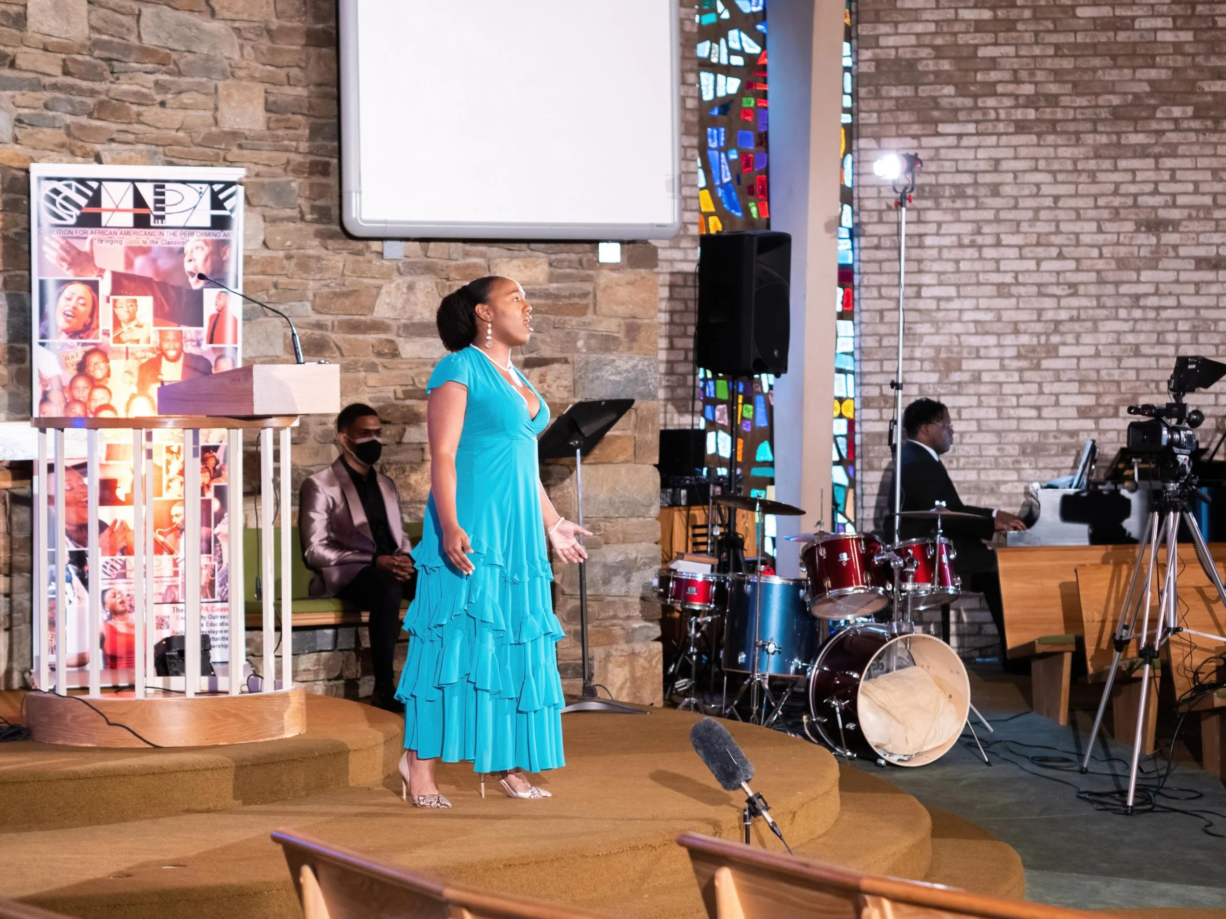 A woman in a flowing blue dress singing on stage in a church or concert hall setting, with a musician playing keyboard, a person sitting behind her wearing a gray blazer and black mask, a drum set, and camera equipment capturing the performance.