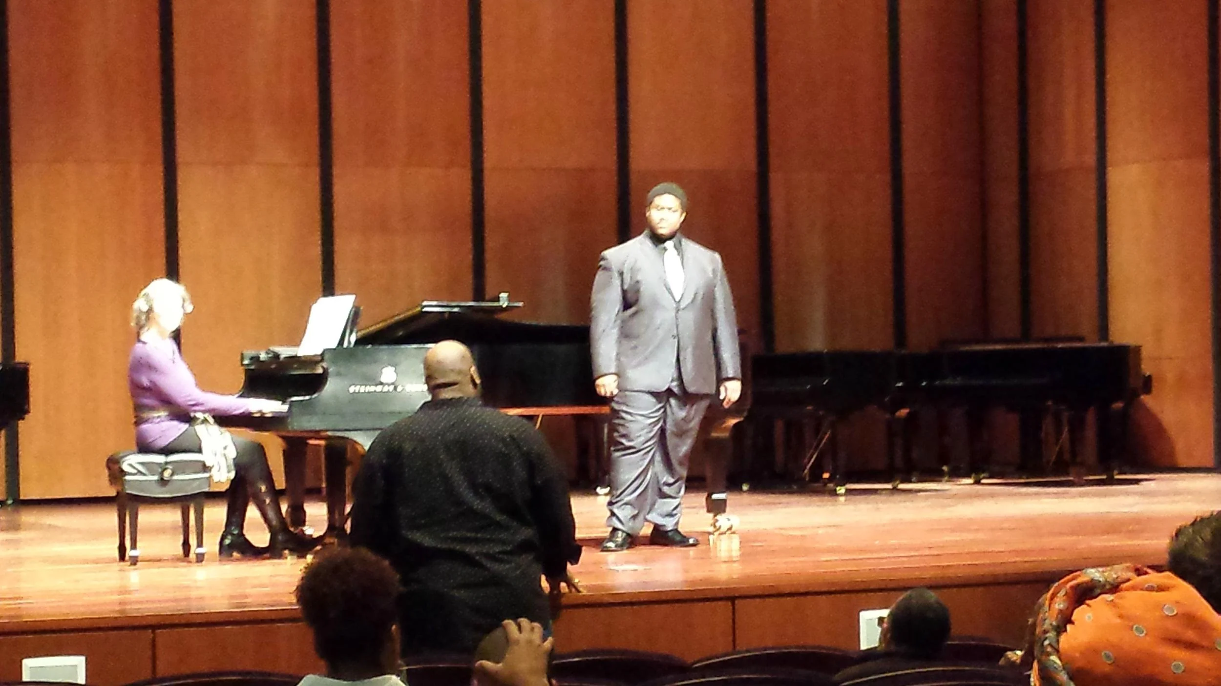 A concert hall with a pianist sitting at a grand piano on the left, a male singer in a gray suit standing in the middle, and an audience watching.