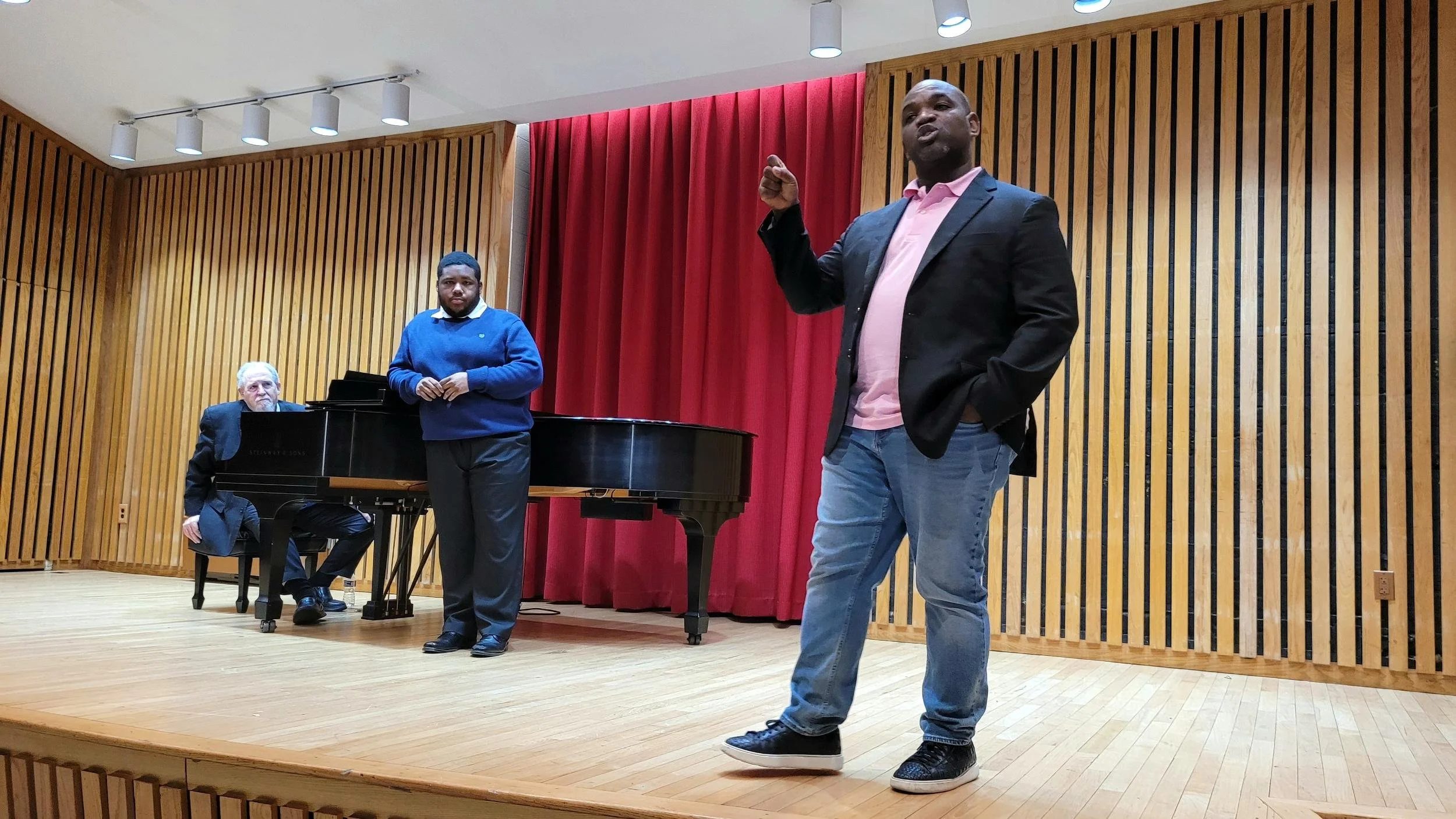 Three men on a stage in a music or performance venue with wooden walls and a red curtain background. One man is sitting at a black grand piano, another man, standing, is dressed casually in a blue sweater and black pants, and the third man, in the fo