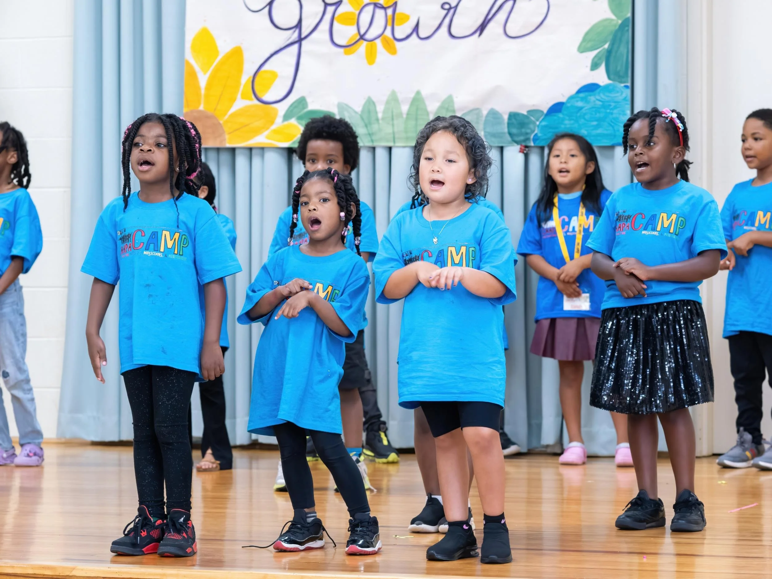 Group of young children singing or performing on stage during a camp event, dressed in matching blue camp t-shirts.
