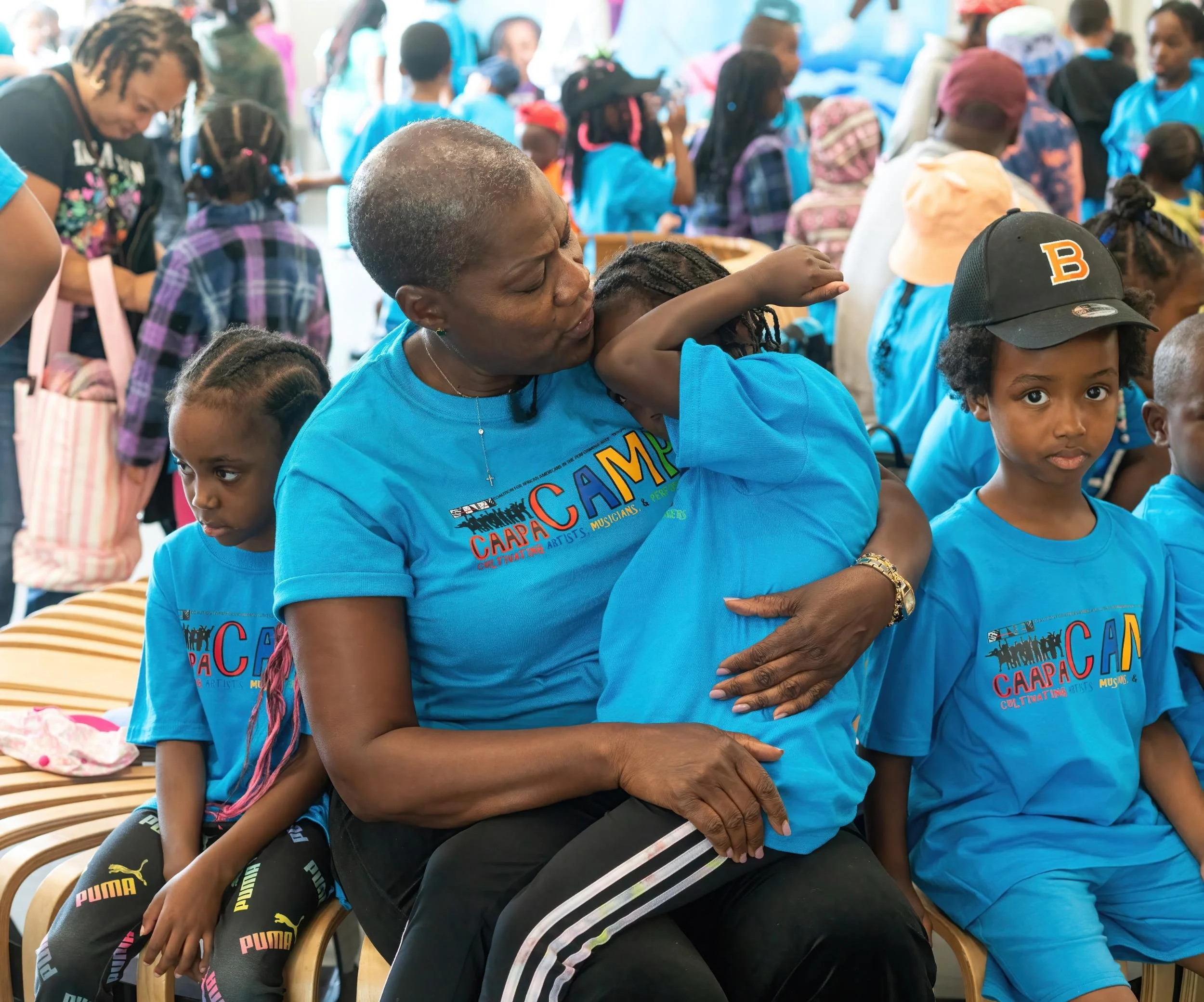 A woman and a young girl share a hug at a crowded event. Both are wearing blue T-shirts with 'CAMP' printed on them, surrounded by children and adults also dressed in blue at an outdoor gathering.