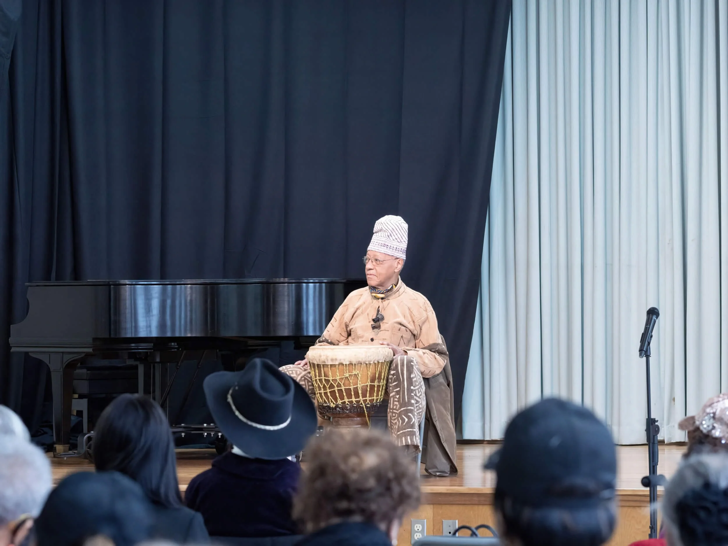 An elderly man wearing traditional African clothing and a striped hat, sitting on a stool and playing a drum on a stage with a black curtain and a piano in the background. An audience is watching the performance.