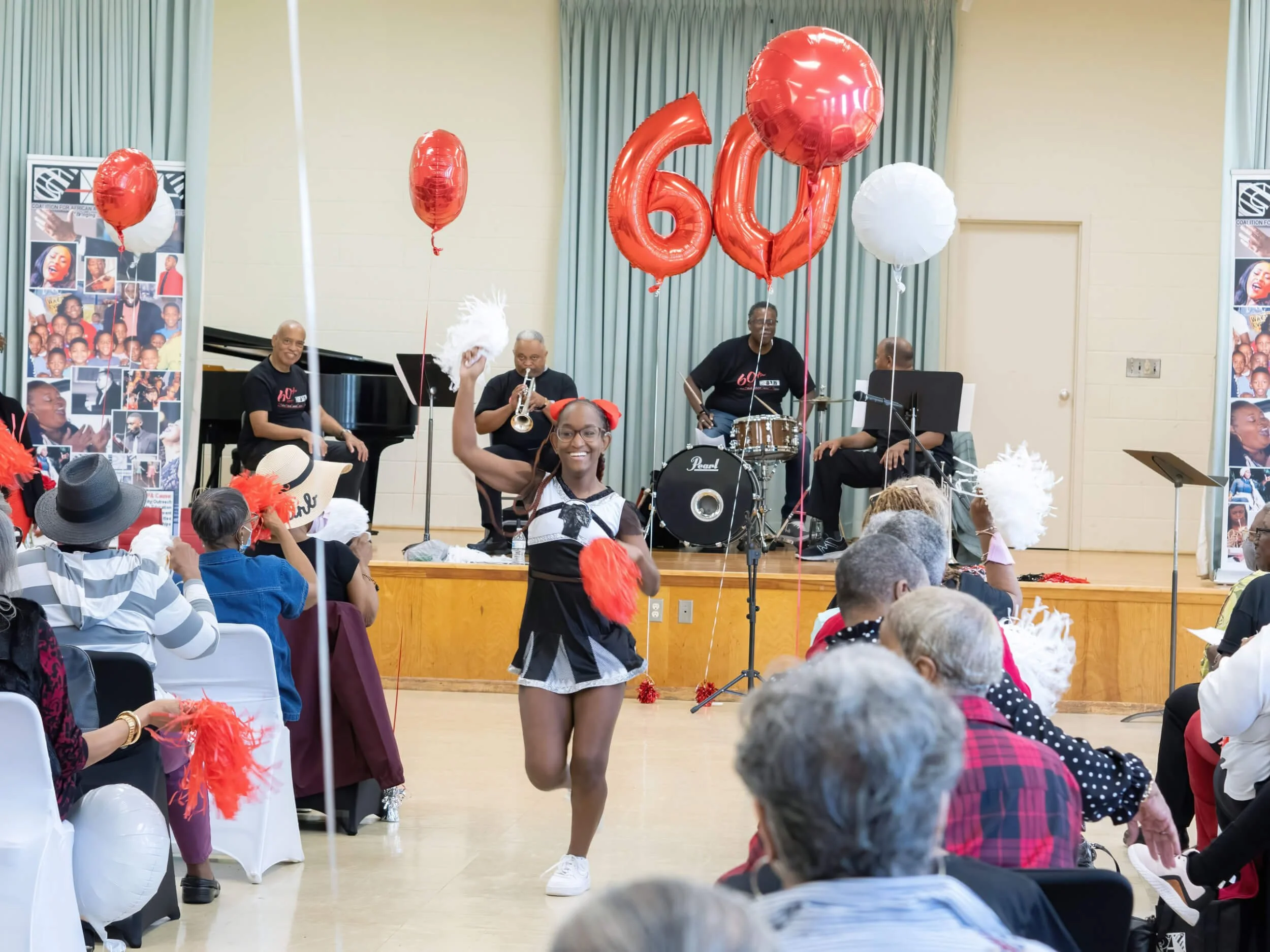 A young girl dances in front of an audience at a celebration event, with a band playing on stage and large red and white balloons that spell out '60' in the background.