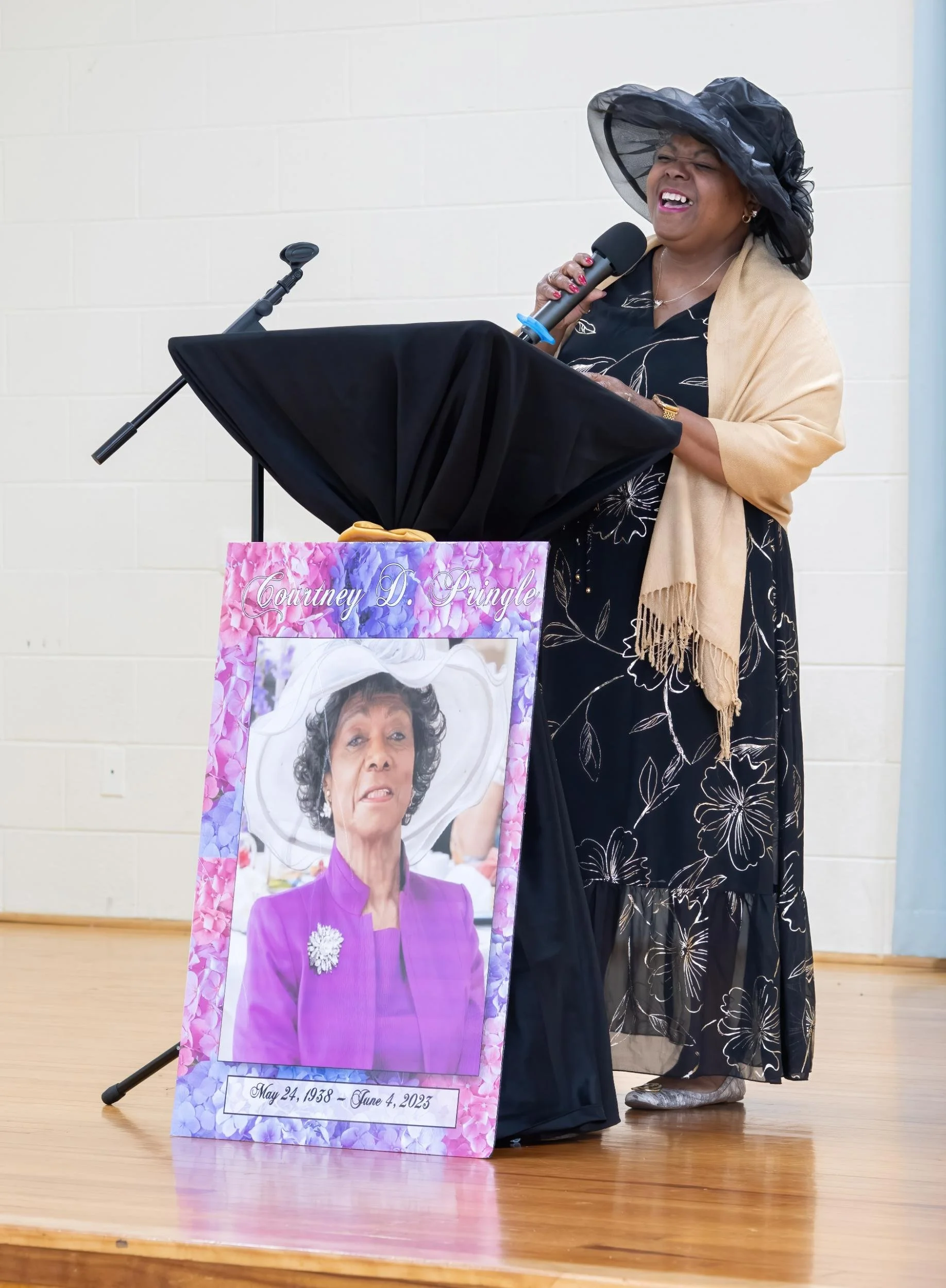 A woman in a floral dress, beige shawl, and large black hat, standing at a podium with a microphone, laughing. A large framed photo of an older woman in purple is on the floor in front of her, with her birth and death dates, May 24, 1938 to June 4, 2