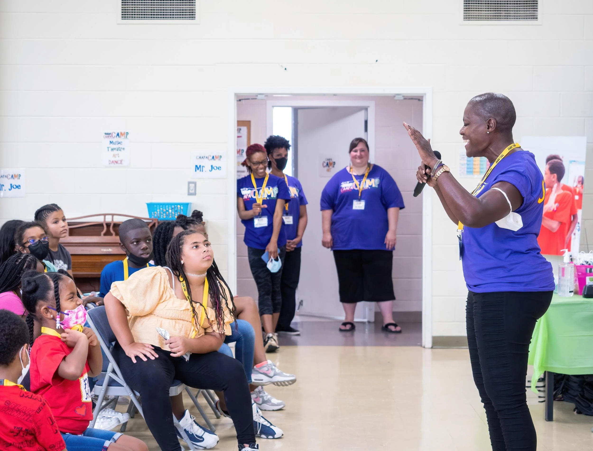 A woman speaking to a group of children seated in a room during a camp activity, with three adults standing in the doorway smiling.