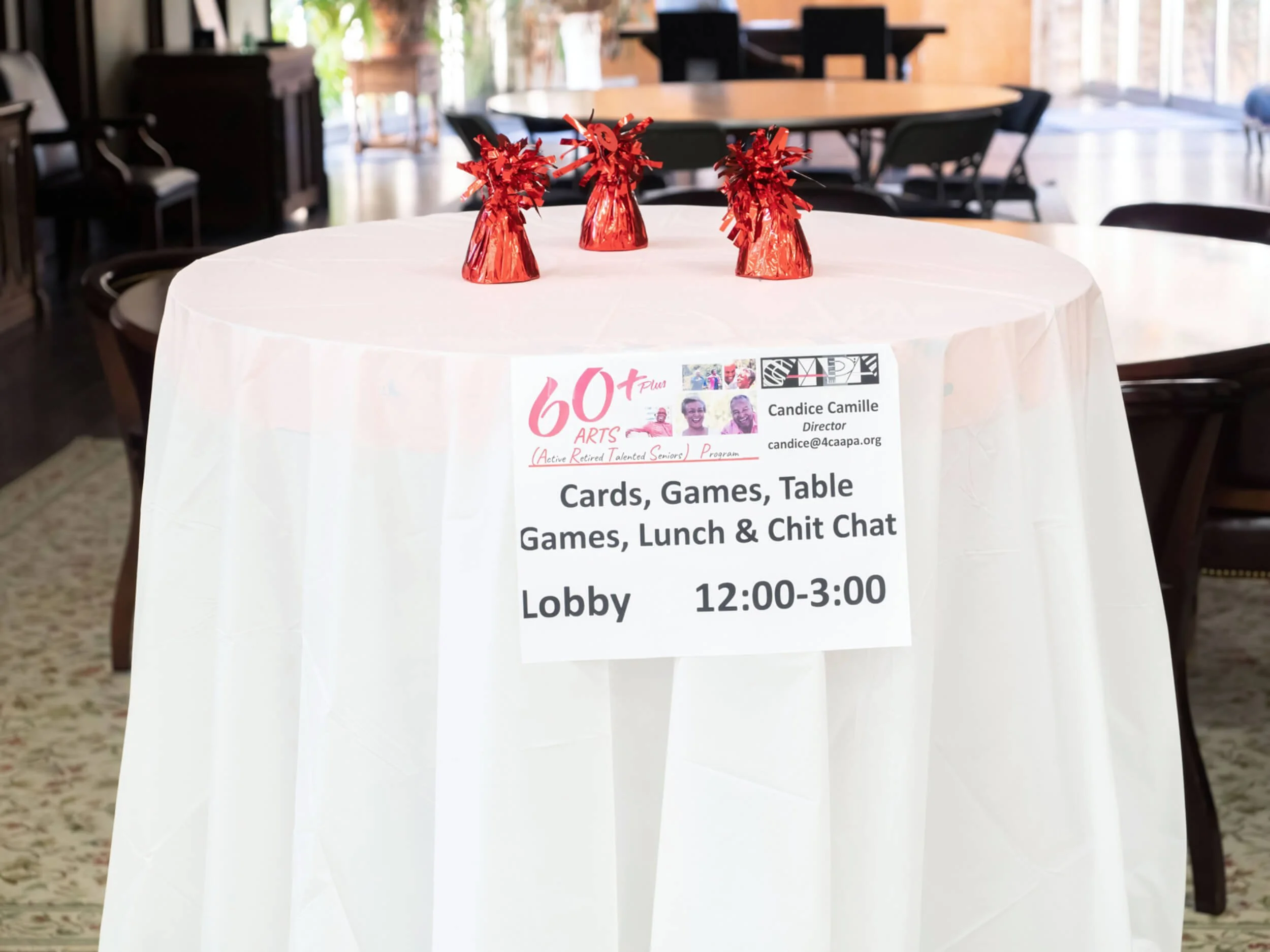A table covered with a white tablecloth inside a brightly lit room, with three small red gift bags with metallic tissue paper on top, and a sign hanging on the front of the table advertising activities for a senior program in the lobby from 12:00 to