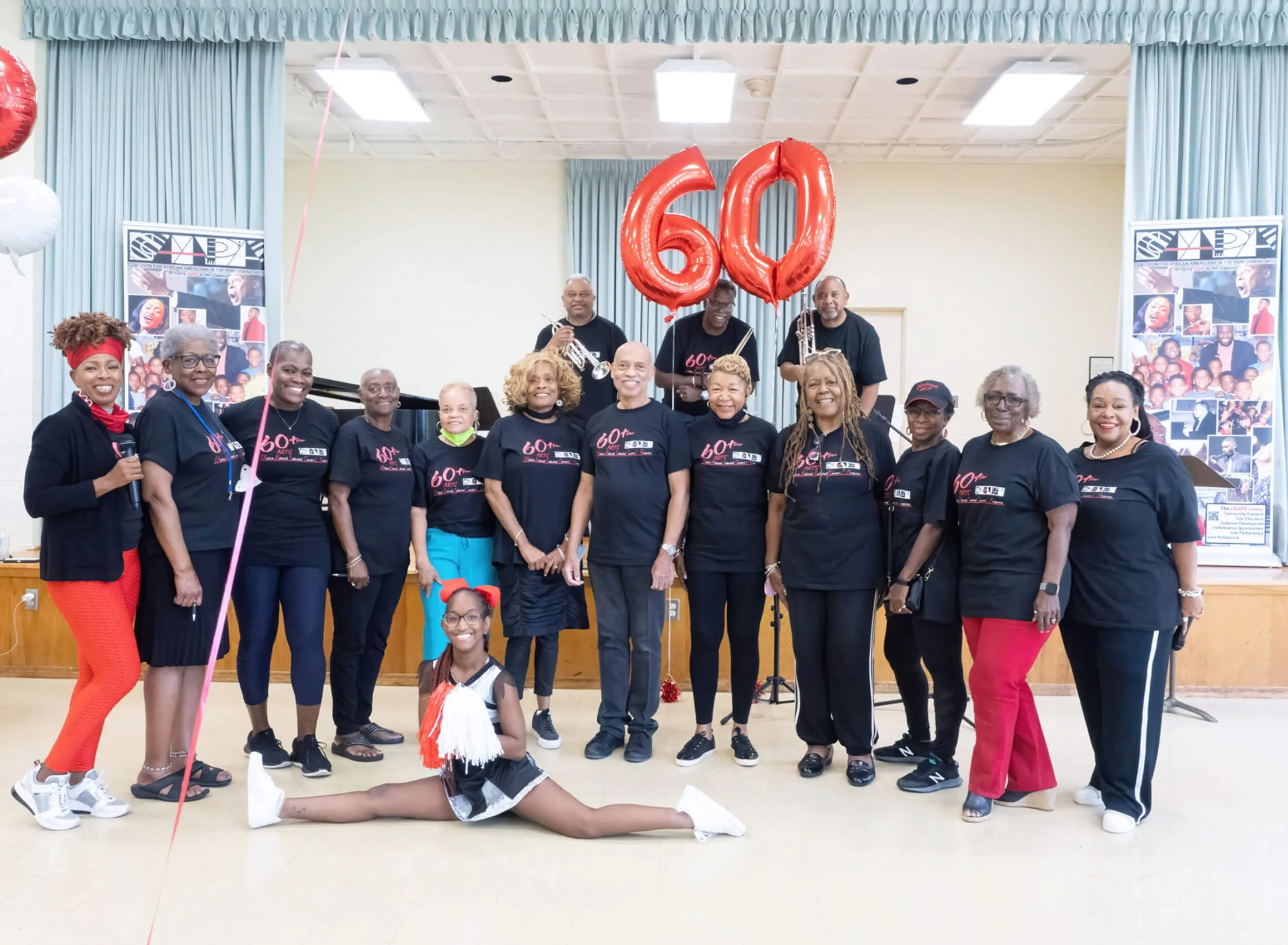 Group of people celebrating 60th anniversary, some holding red balloons shaped as the number 60, one person performs a split in front with a pom-pom in hand, all wearing matching black T-shirts, in a decorated indoor setting.