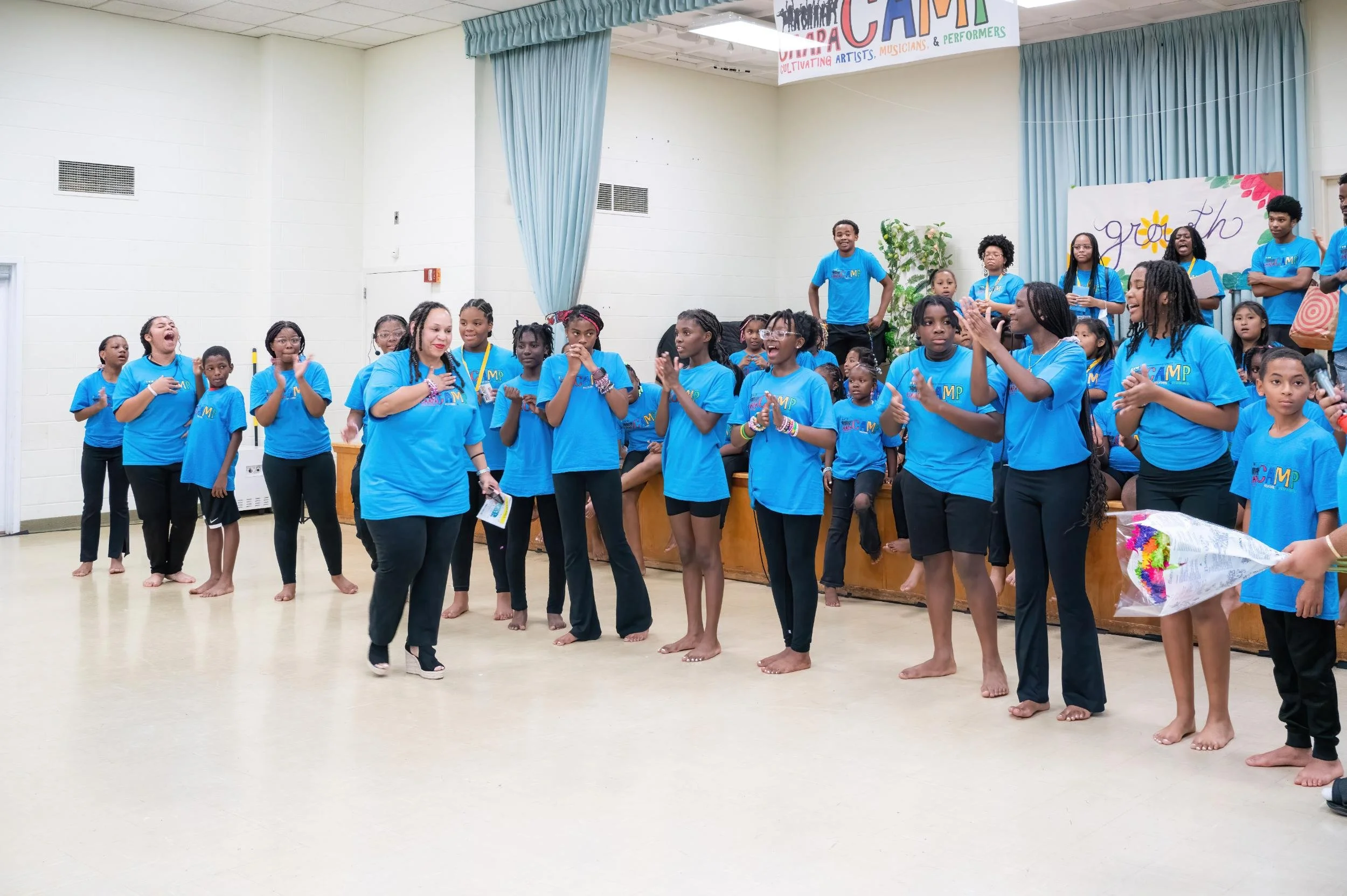 Group of children and adults in matching blue T-shirts at a graduation or award ceremony, standing barefoot in a large room with white walls, blue curtains, and banners that say 'Graduation' and 'Camp'.