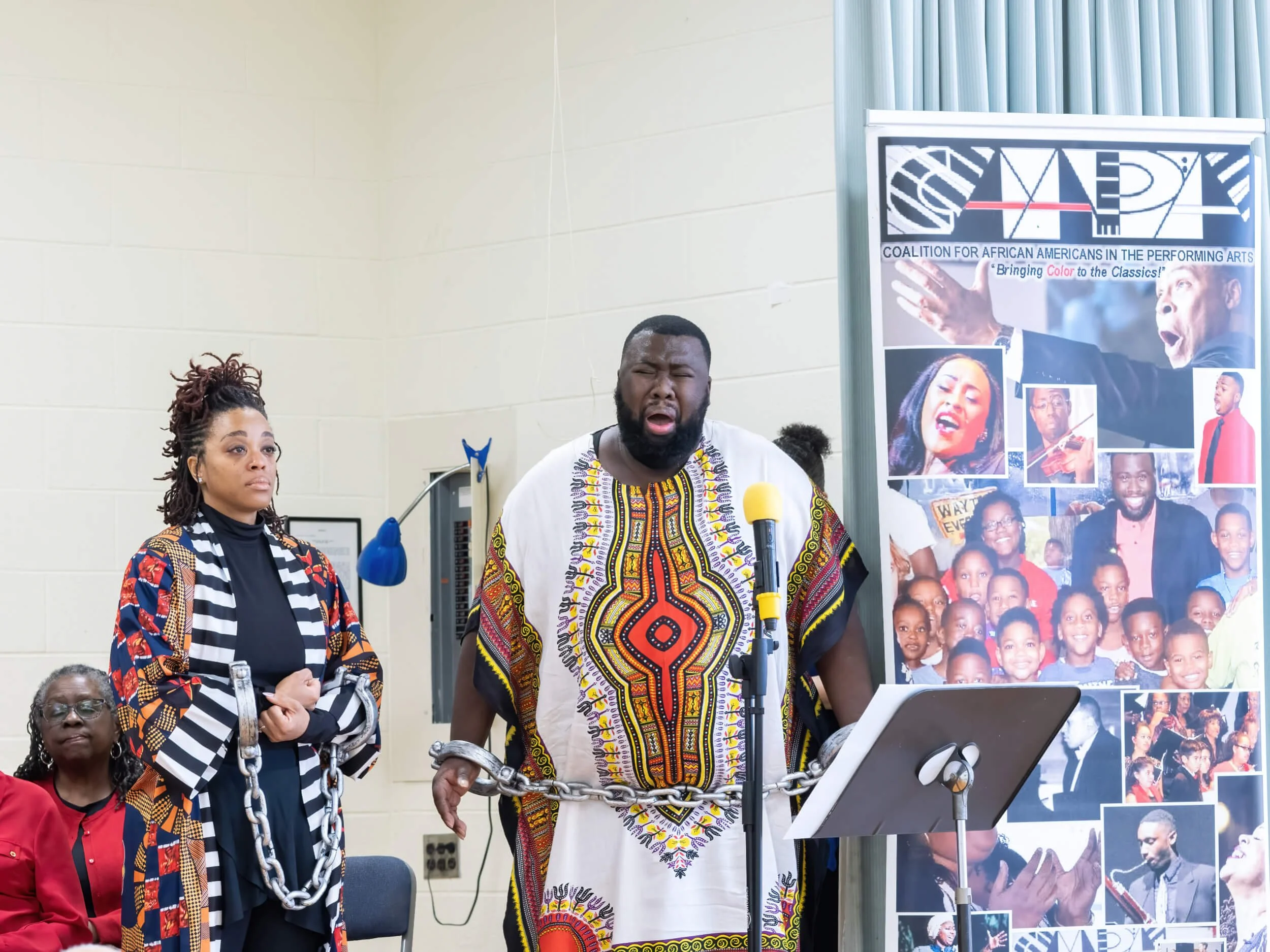 A man in traditional African attire is speaking or singing into a microphone, with two women standing nearby, one with a serious expression and the other in red, at an indoor event for the Coalition for African Americans in the Performing Arts. A lar