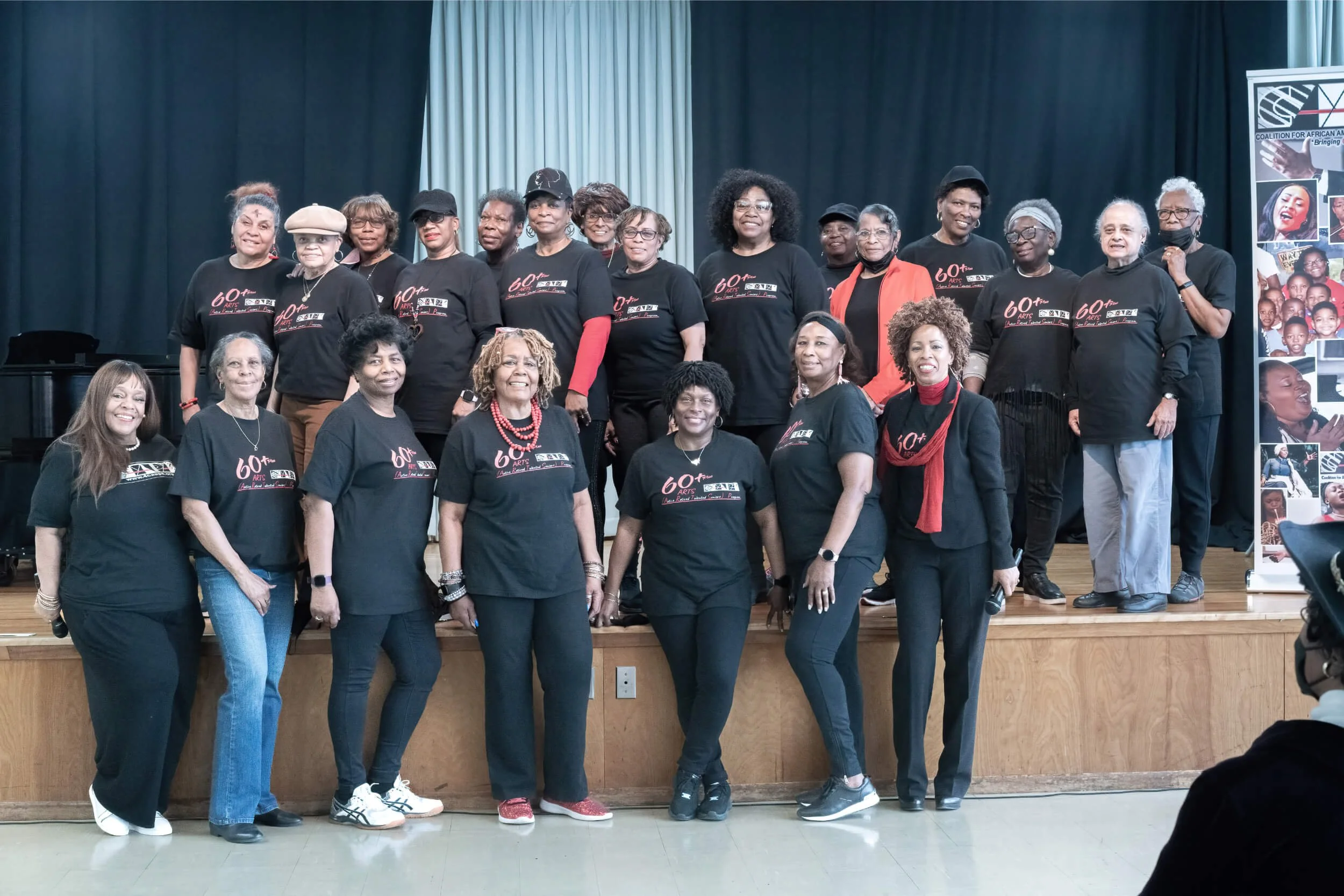Group of women on stage wearing matching black T-shirts with red and white text, celebrating an event, with a banner on the side featuring photos and text.