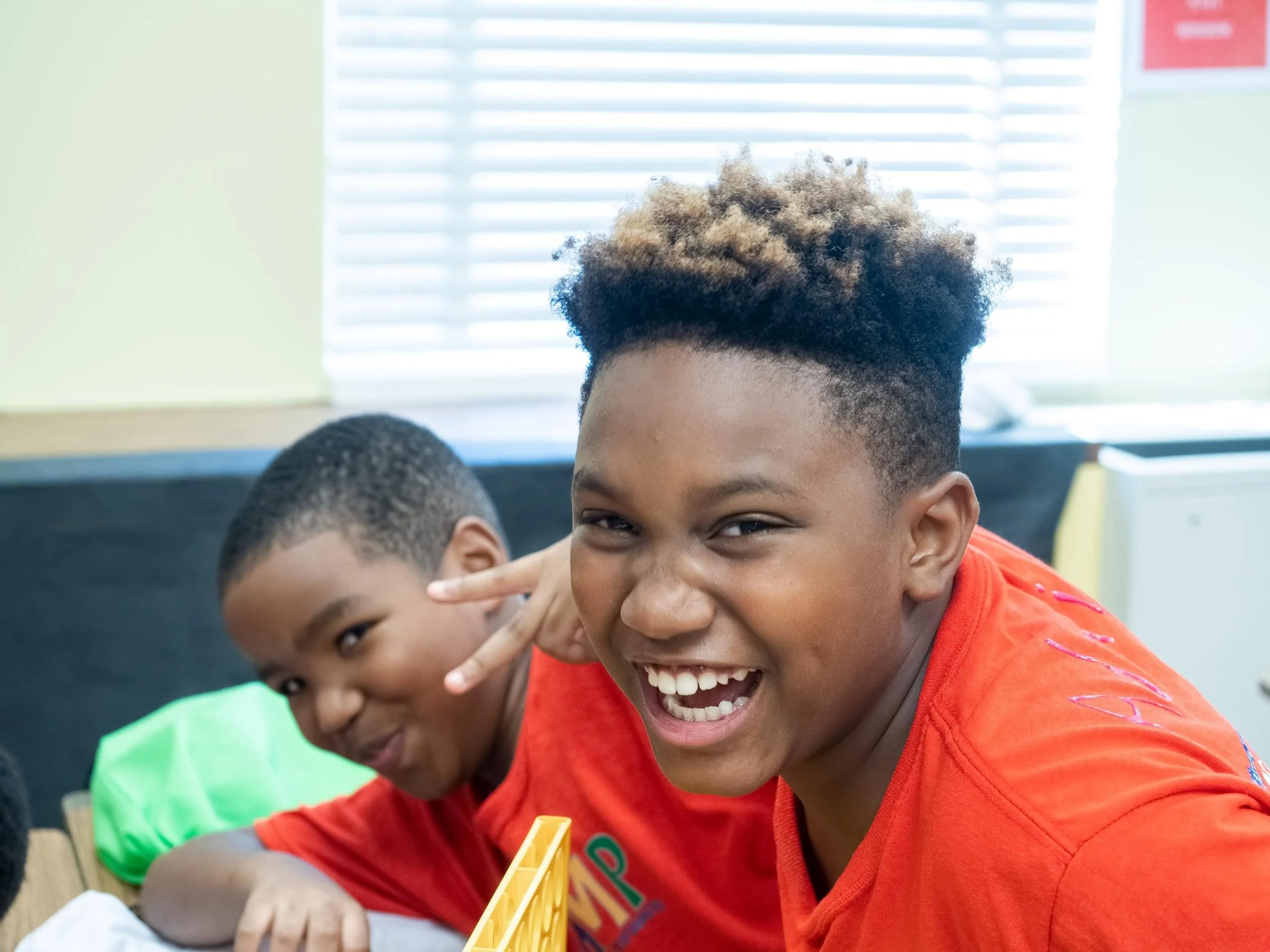 Two boys smiling and making playful gestures in a classroom, one wearing a bright orange shirt and the other with a green backpack nearby.
