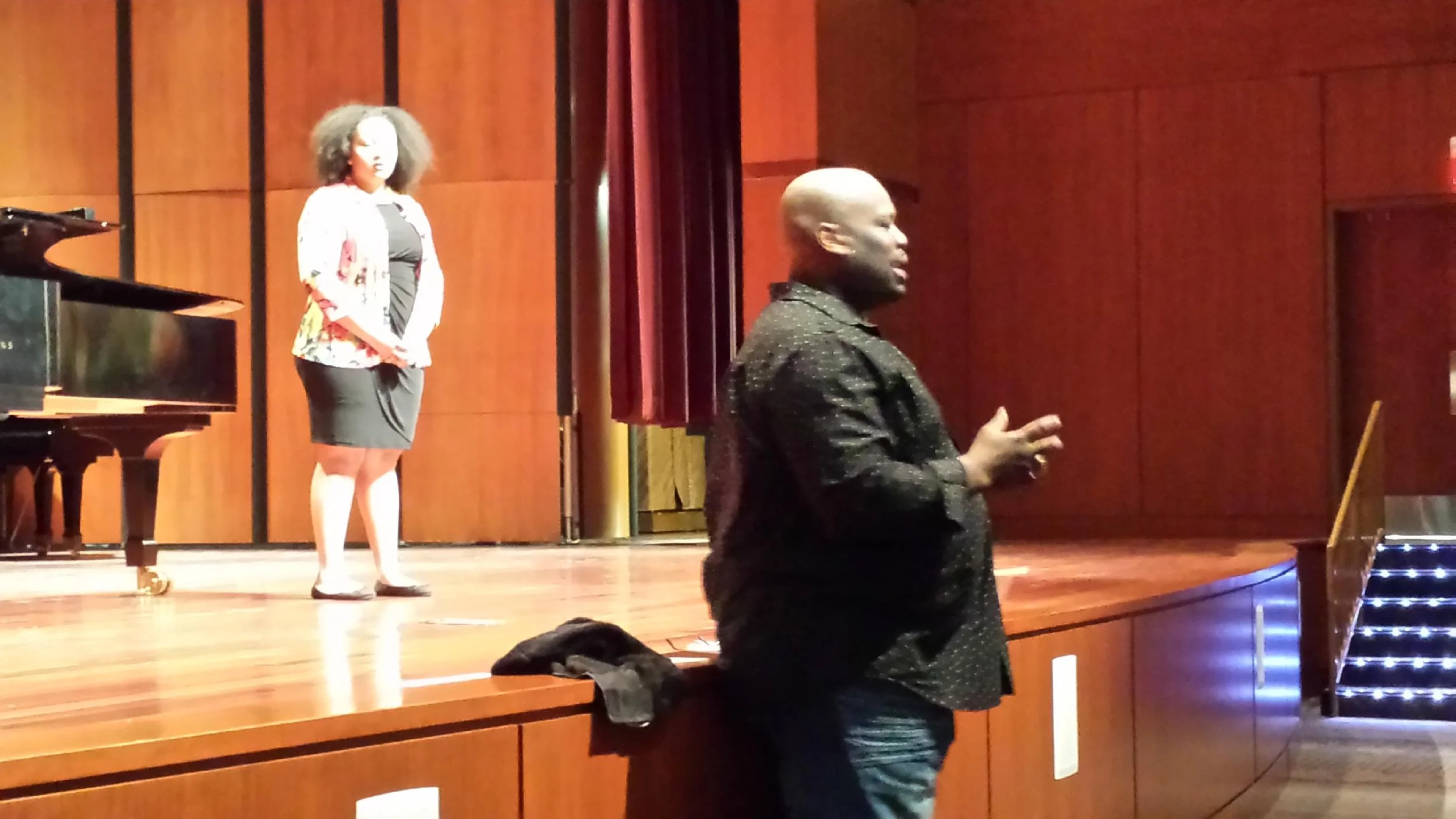 A man standing at the edge of a stage in a wood-paneled auditorium, talking or giving a presentation. A woman is standing on the stage in the background near a piano, listening.