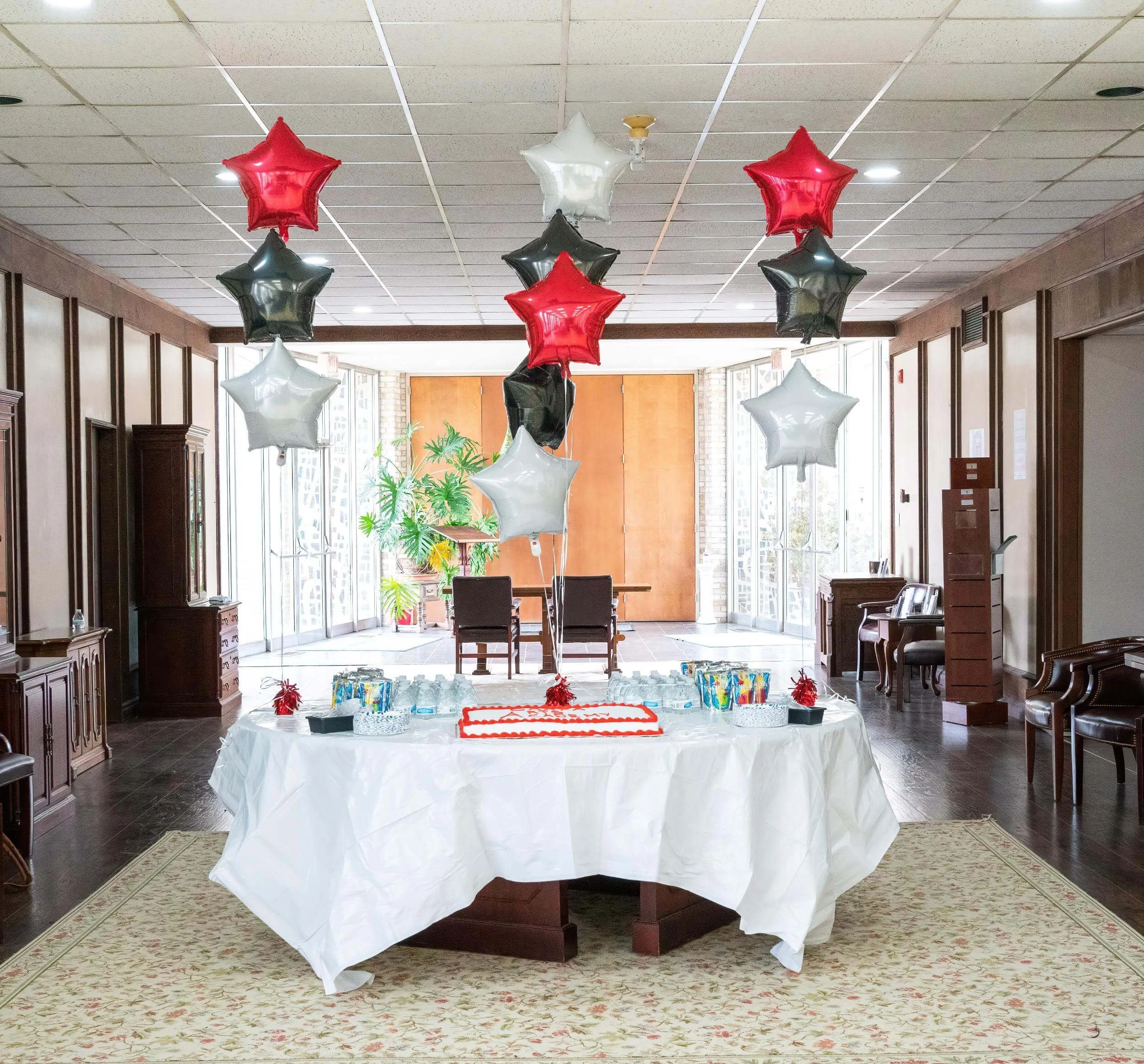 Decorative balloons in red, black, and white hang above a table with a cake and drinks in a well-lit room.