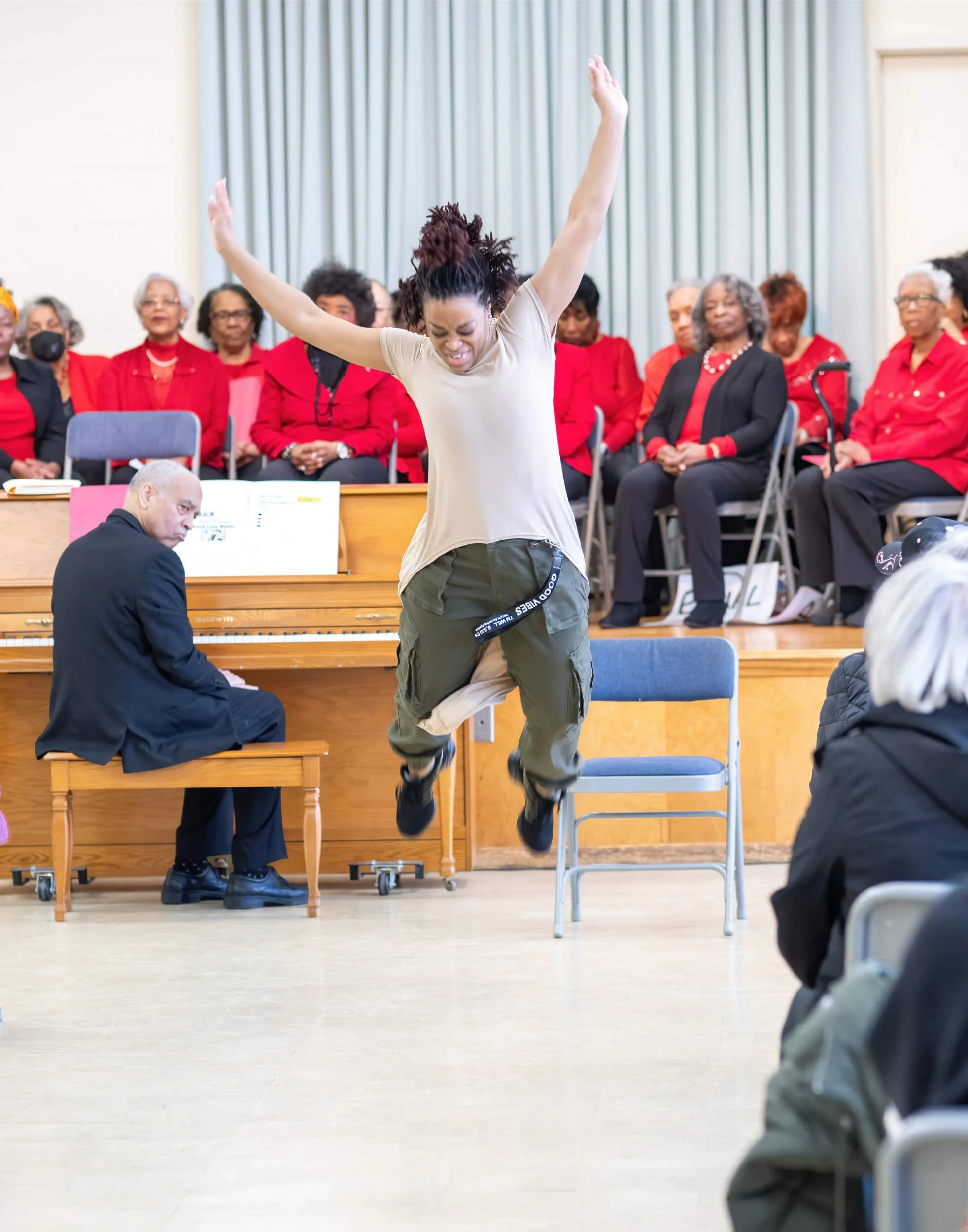 A woman jumping in front of a seated choir, with an older man playing the piano nearby, during a performance or event.