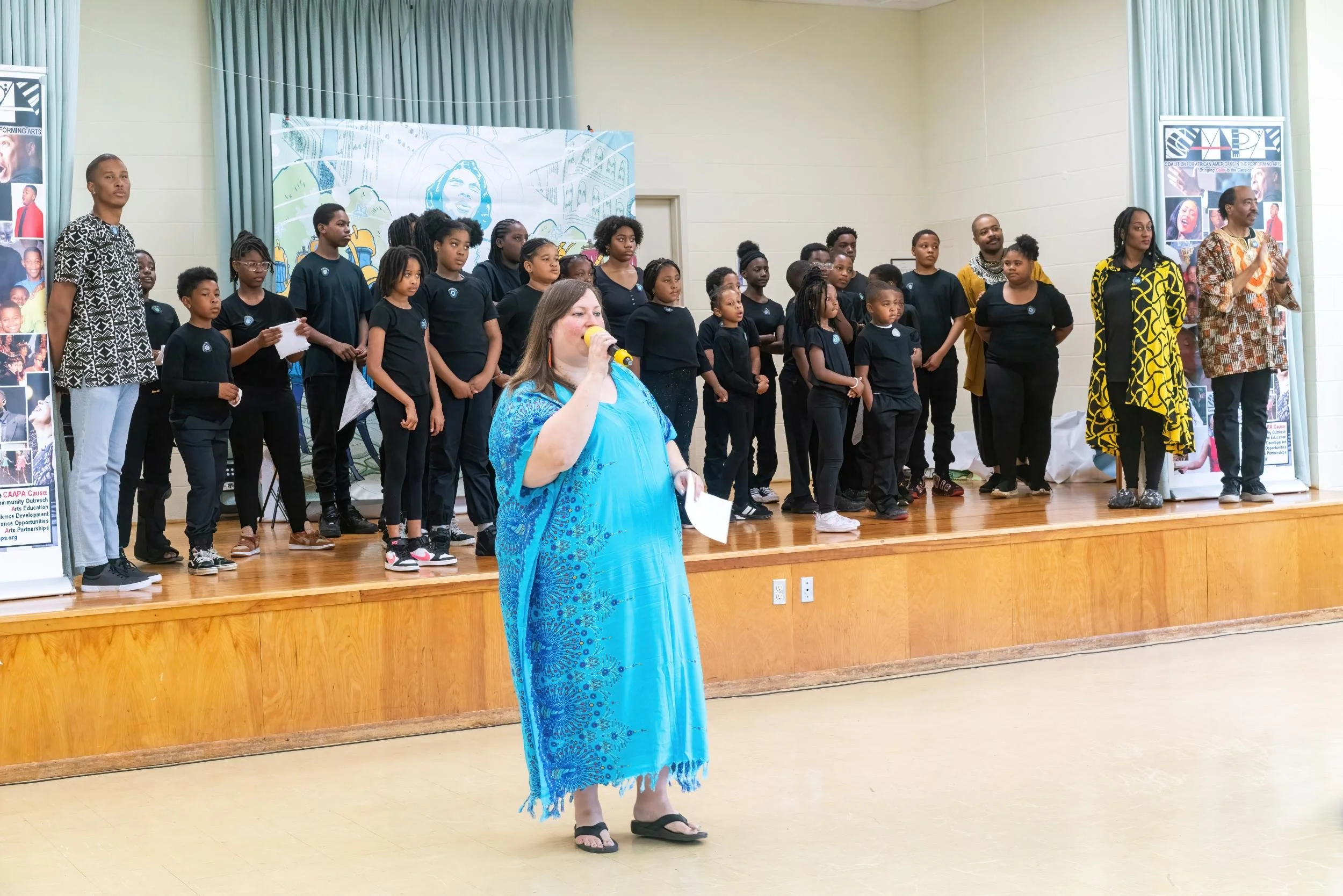 A group of children and adults standing on a stage during a performance or event, with a woman in a blue dress speaking into a microphone in front of them.