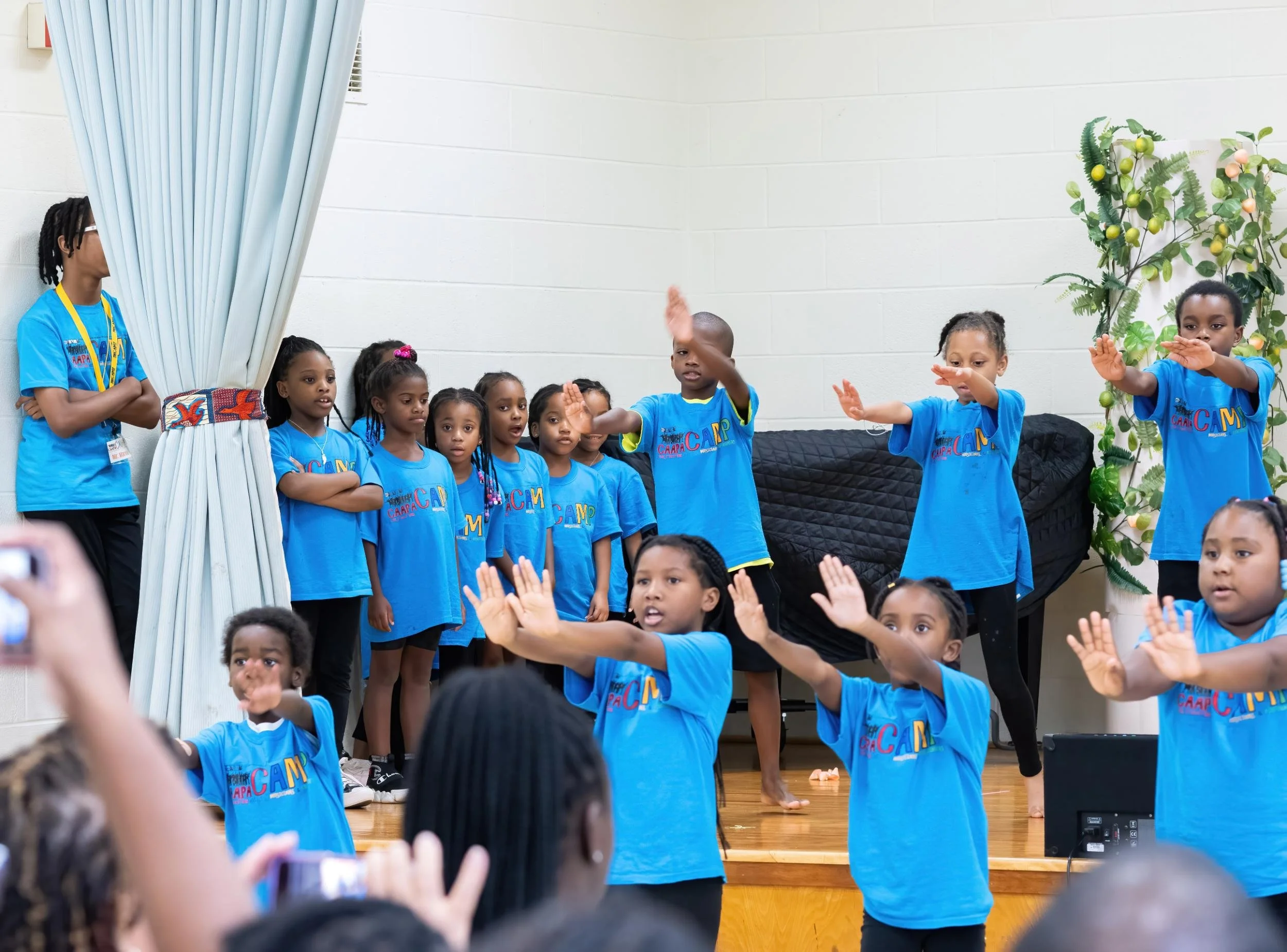 Children performing a dance on stage, some with arms extended, wearing matching blue T-shirts with 'CAMP' printed on them. An audience is visible in the foreground.