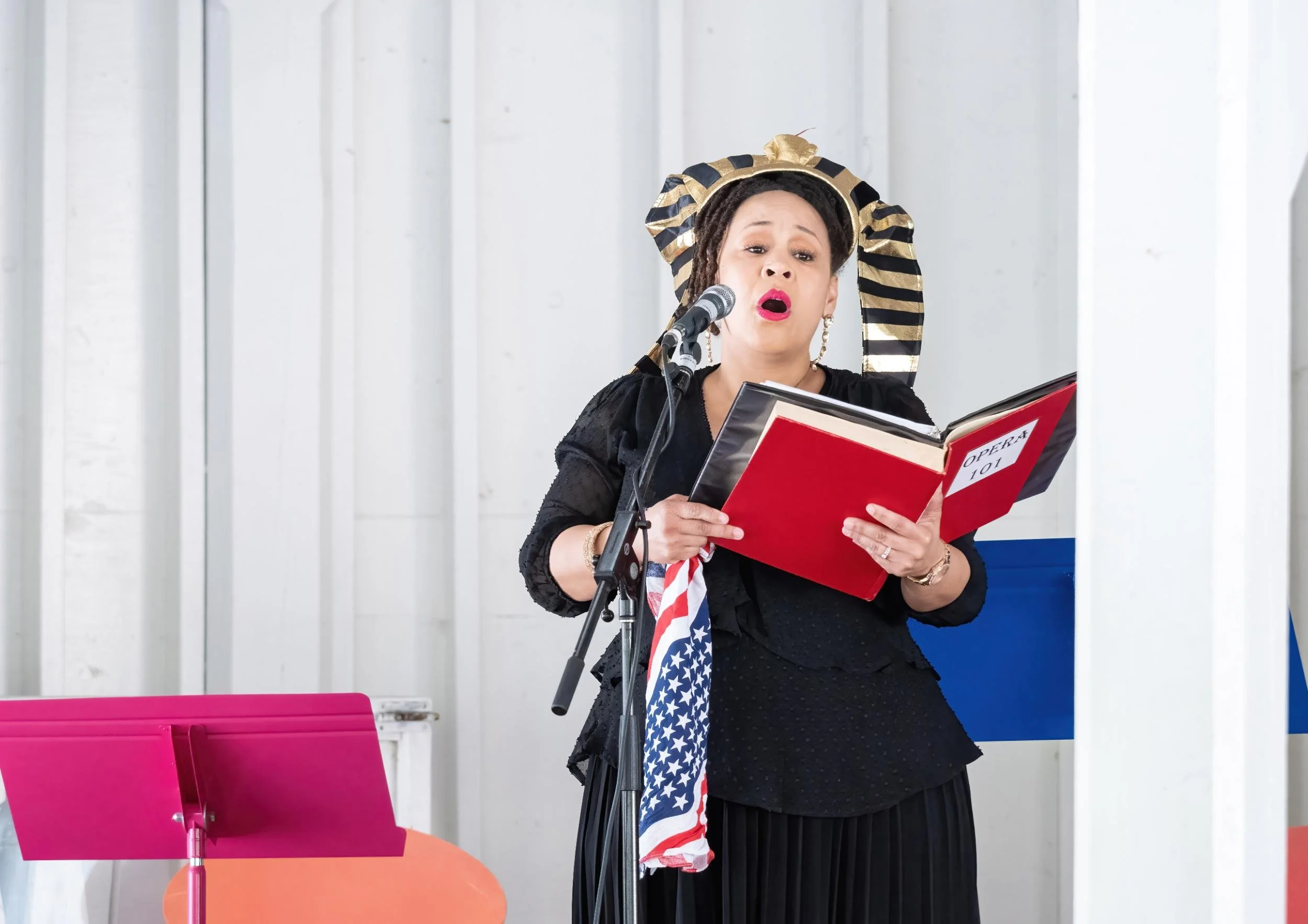 A woman dressed as a singer in black, with a gold and black headscarf, singing into a microphone and reading from a red operatic songbook labeled 'Opera 101'.