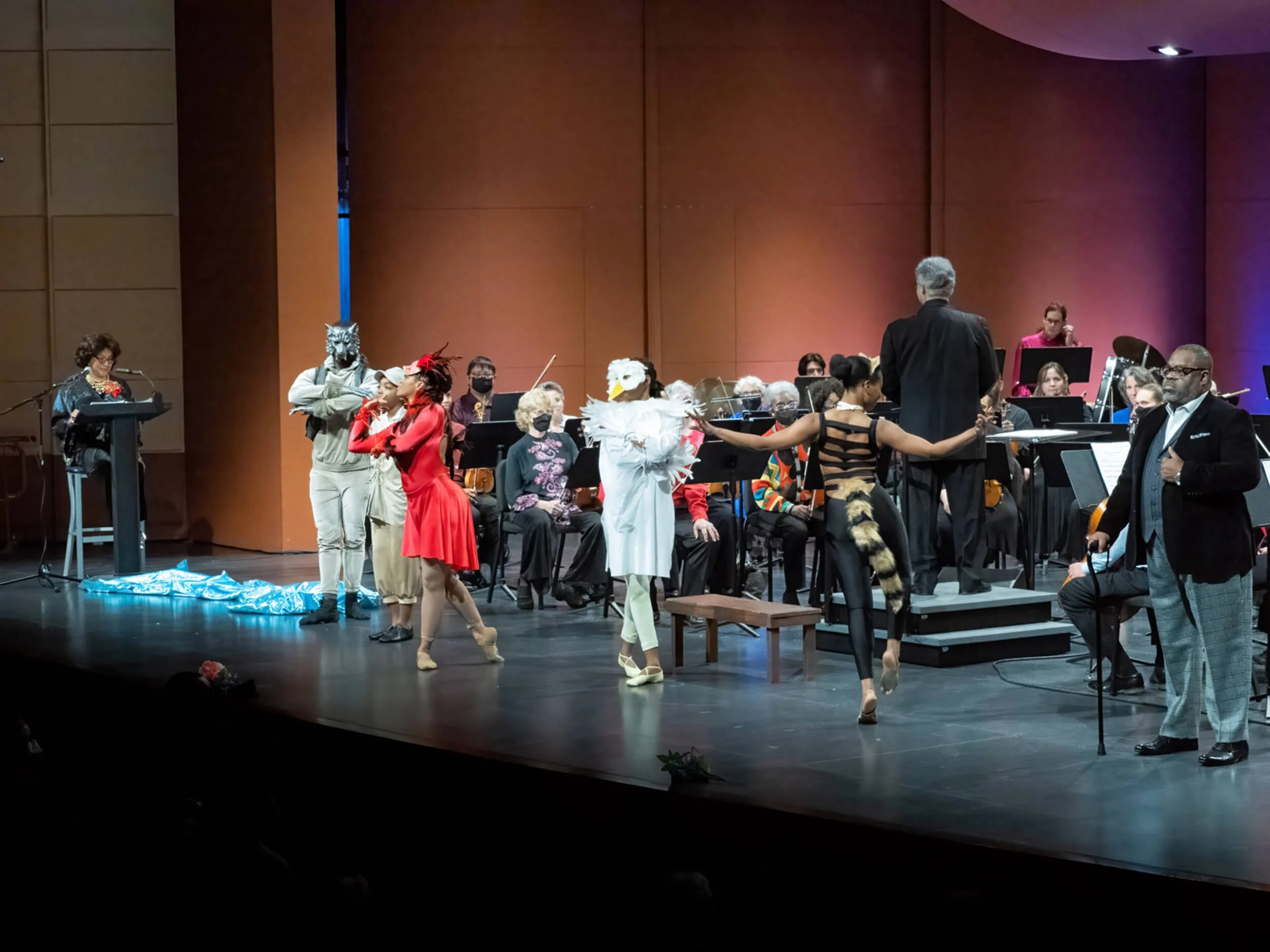 Theater stage with a conductor, musicians, and performers in costumes, some with animal masks, in a concert hall with lighting.