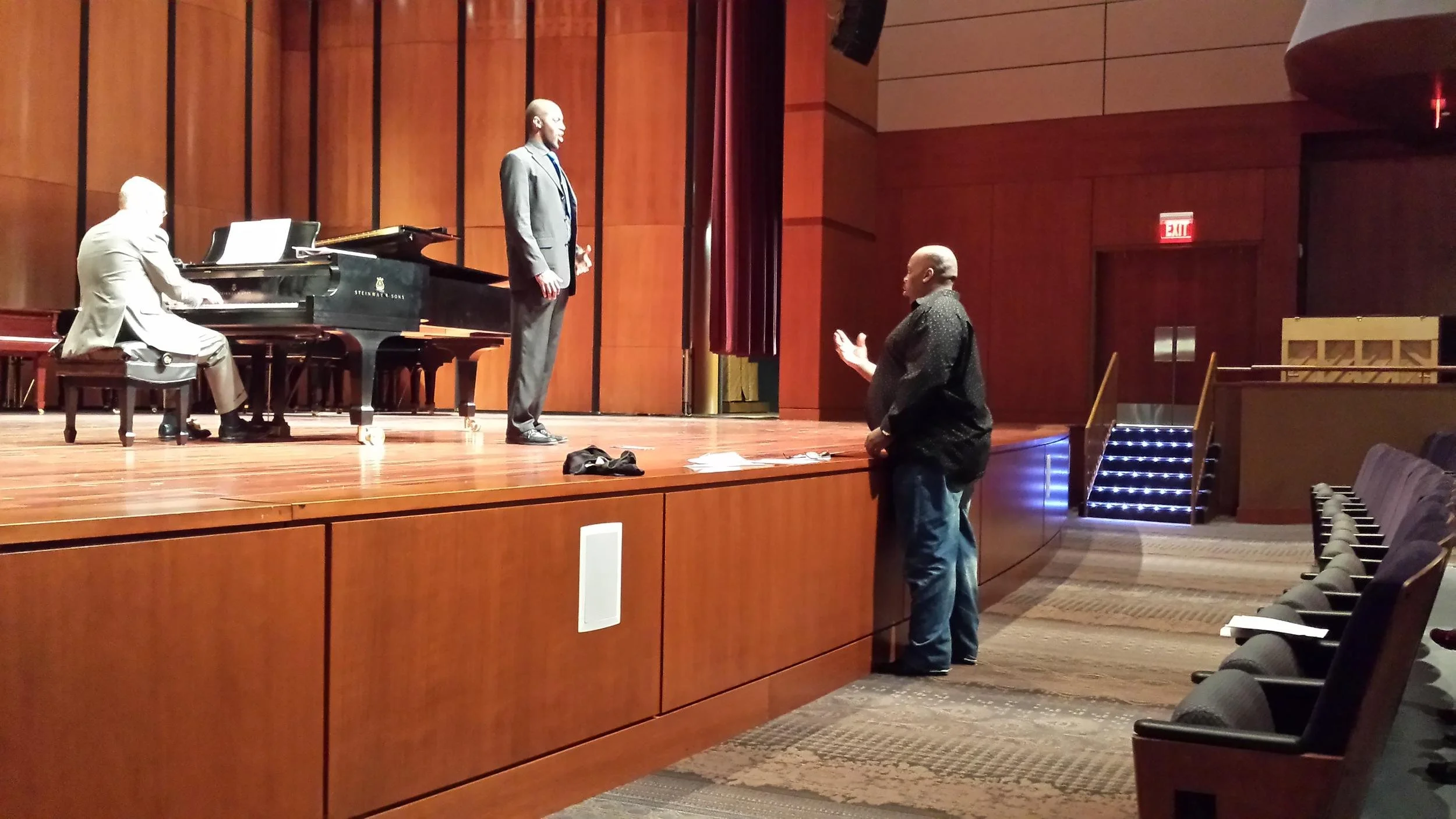 A man kneeling on the floor facing a man standing on stage, with a pianist playing a grand piano nearby. The setting appears to be an auditorium or theater with wood-paneled walls and an exit door with stairs leading to another level.