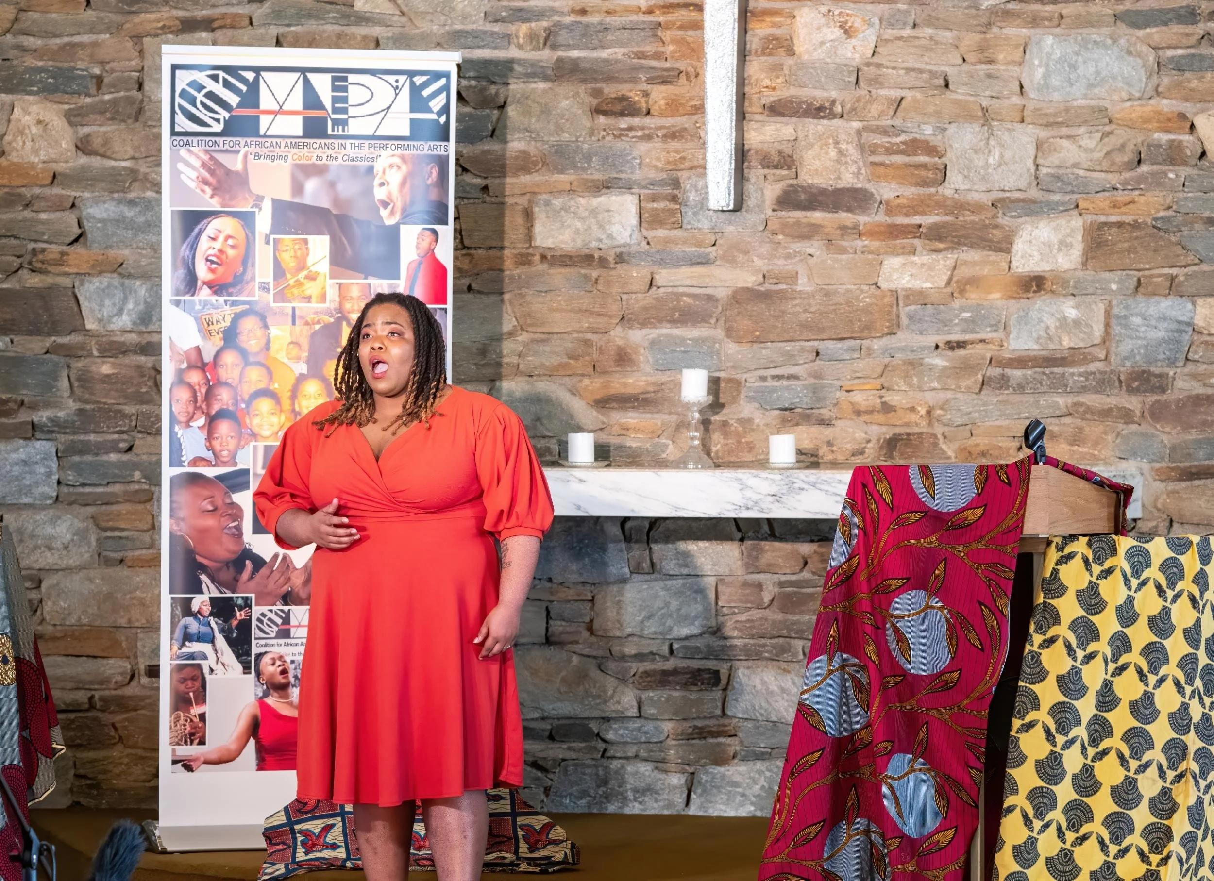 A woman in a red dress singing or speaking on stage, with a stone wall and a banner promoting African American performing arts in the background.