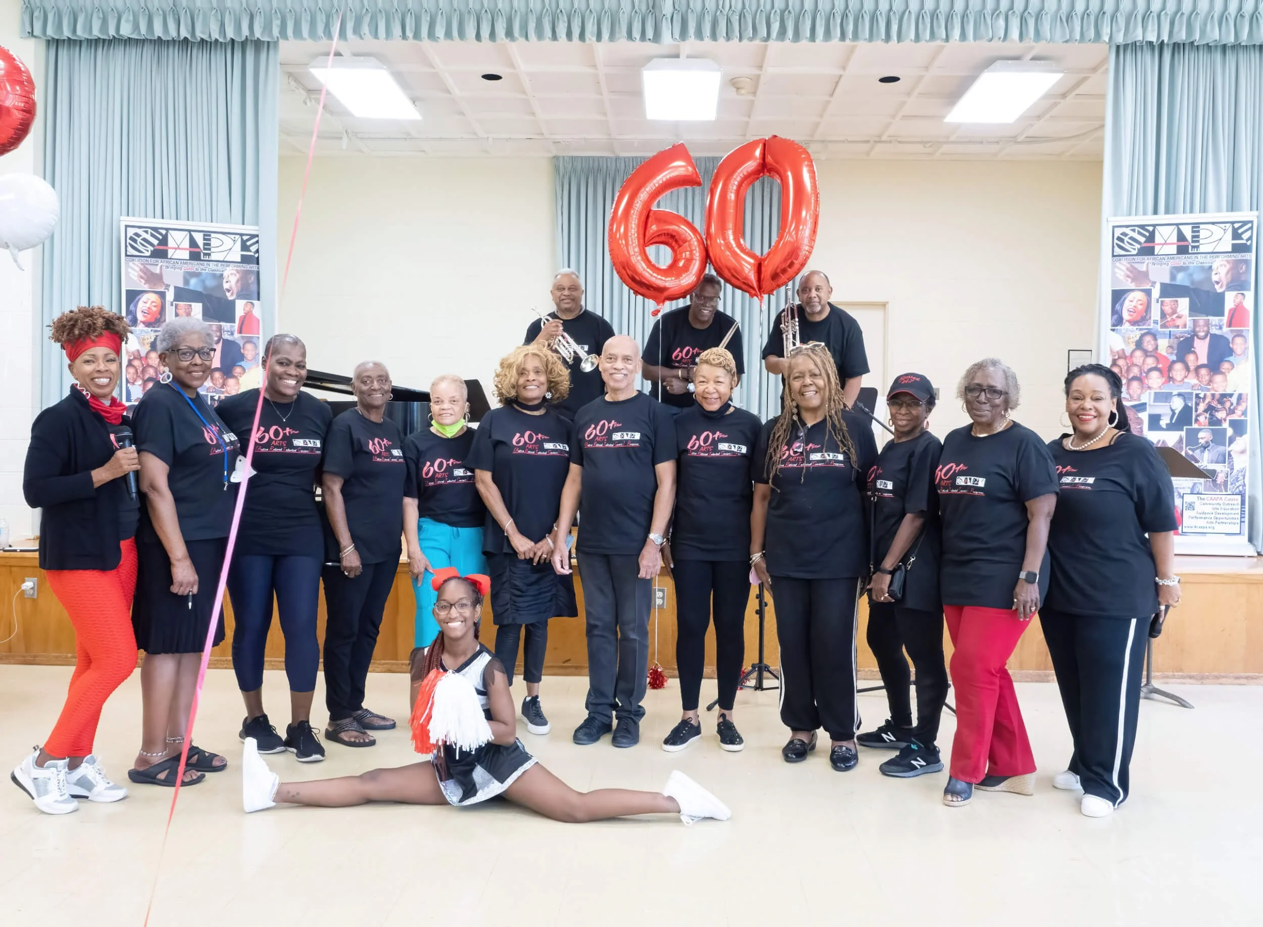 A group of people celebrating a 60th anniversary, with some holding red balloons shaped as the numbers 60, in a decorated room, with a cheerleader girl in front.