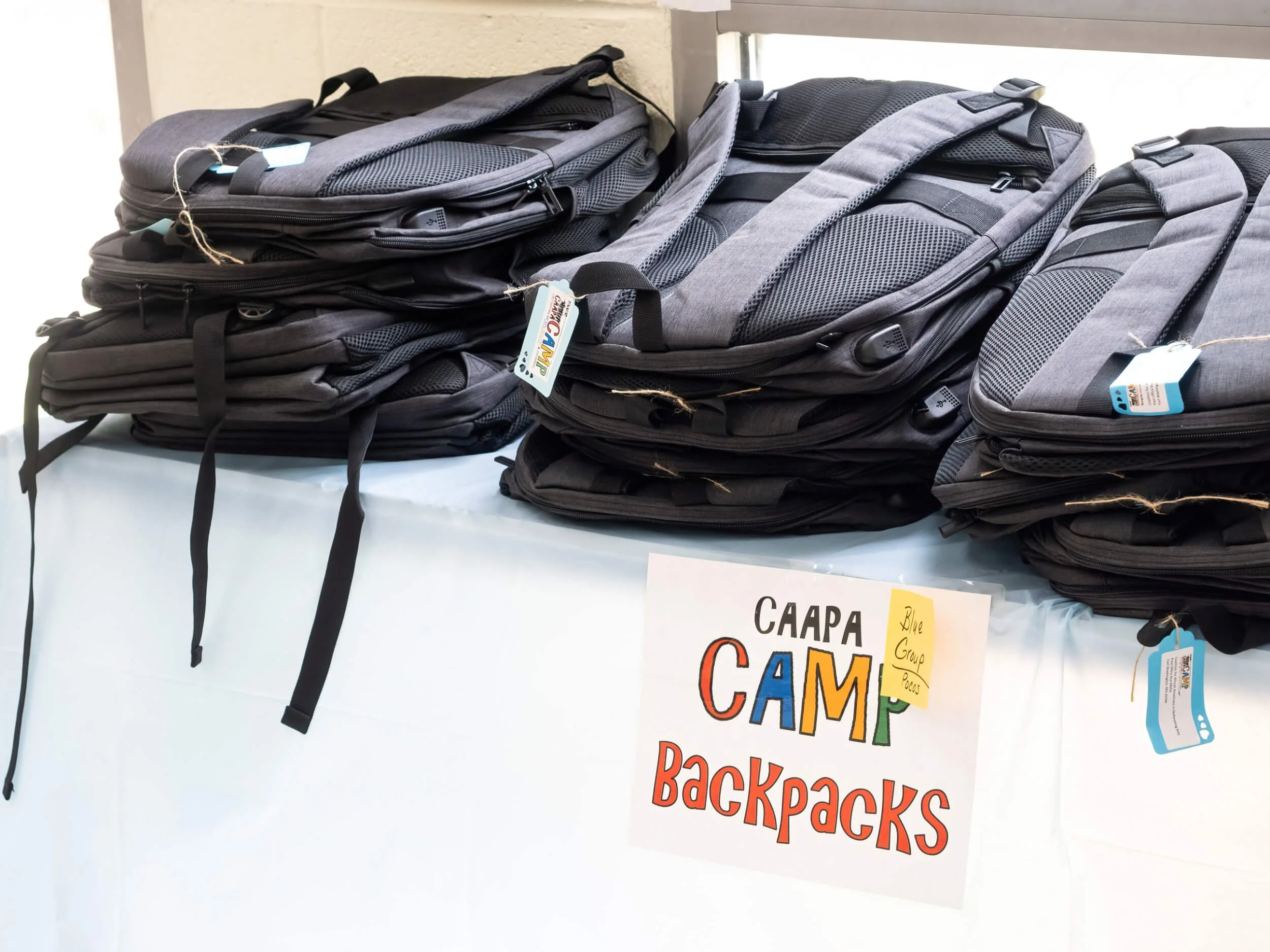 Stacks of gray and black backpacks on a table with a sign that reads "CAMP Backpacks" and a note that says "Blue Group Packs".