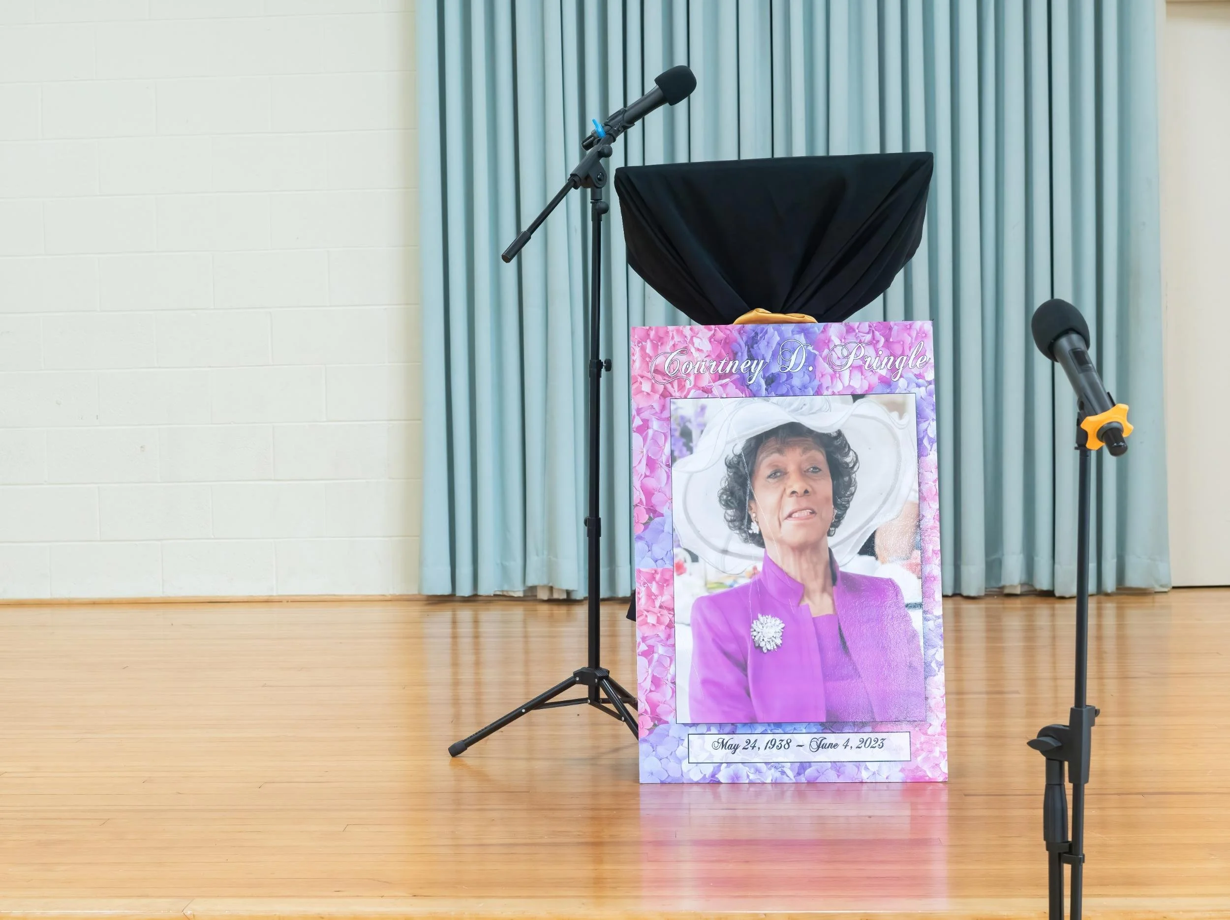 A memorial display with a large framed photo of an elderly woman wearing a white hat and purple outfit, surrounded by pink and purple flowers, with her name, Courtney D. Pringle, and birth and death dates, May 24, 1938 - June 4, 2023, written at the