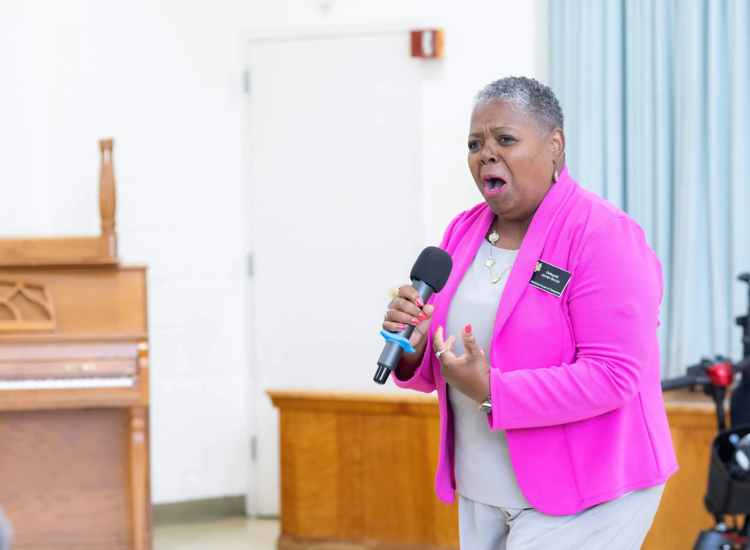 An older woman with short gray hair wearing a pink blazer and jewelry speaking into a microphone in a room with a wooden piano and a white door.