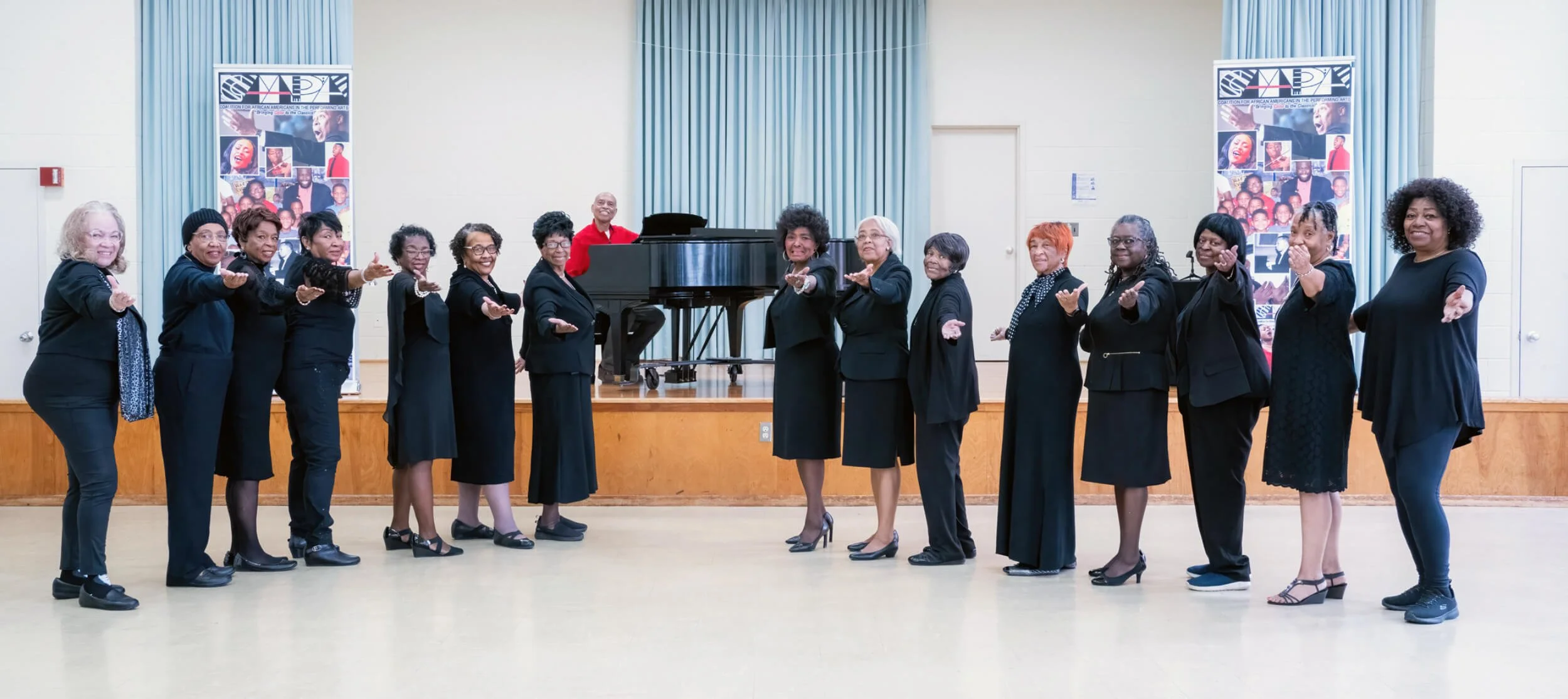 A group of women's choir members standing in a line on stage, posing with arms extended forward, with a man playing piano behind them. They are dressed in black or dark clothing, with two banners with photos and the acronym 'SAGE' in the background.