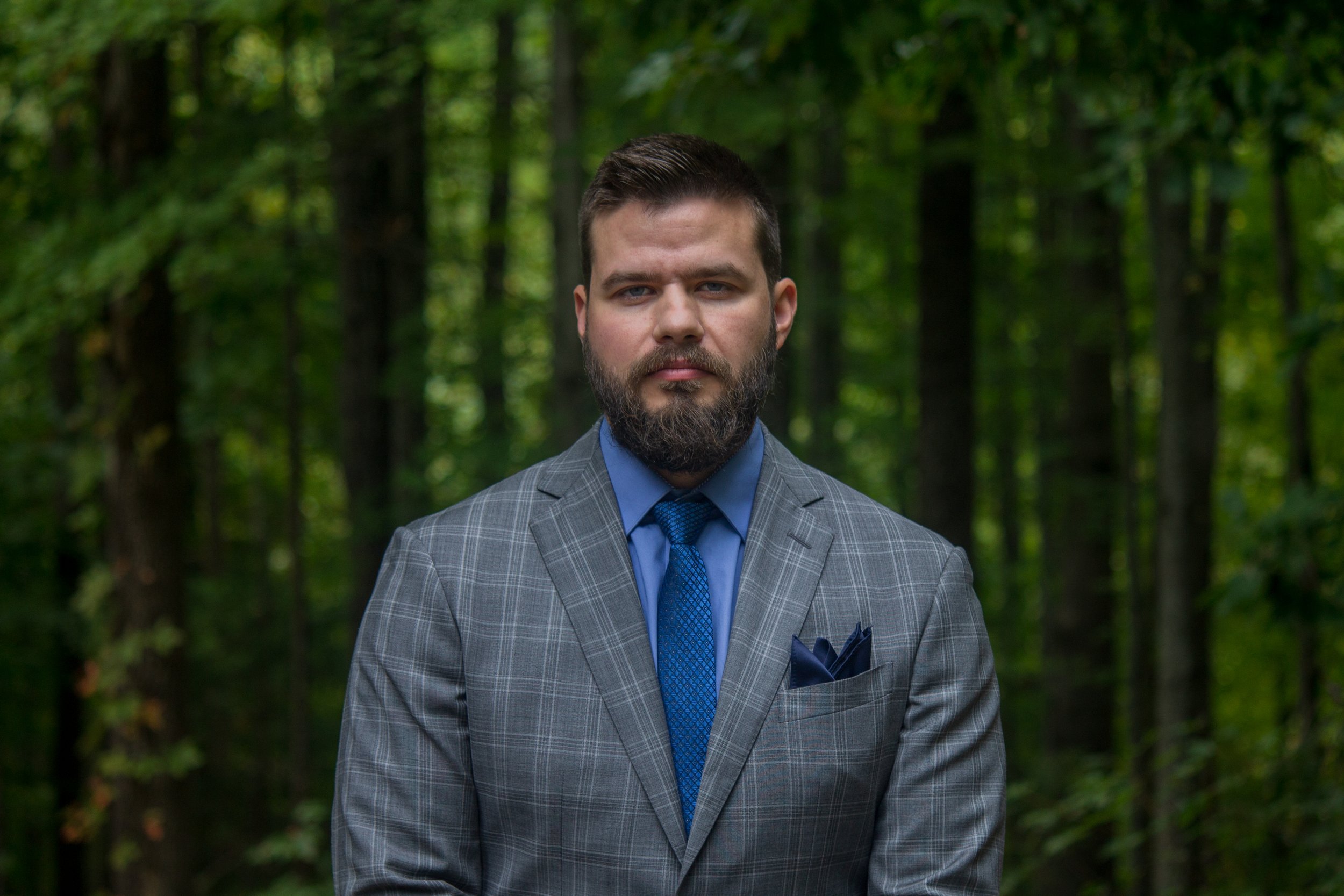 A man, the founder, with a beard wearing a gray checkered suit, blue shirt, and blue tie standing outdoors in a forest.