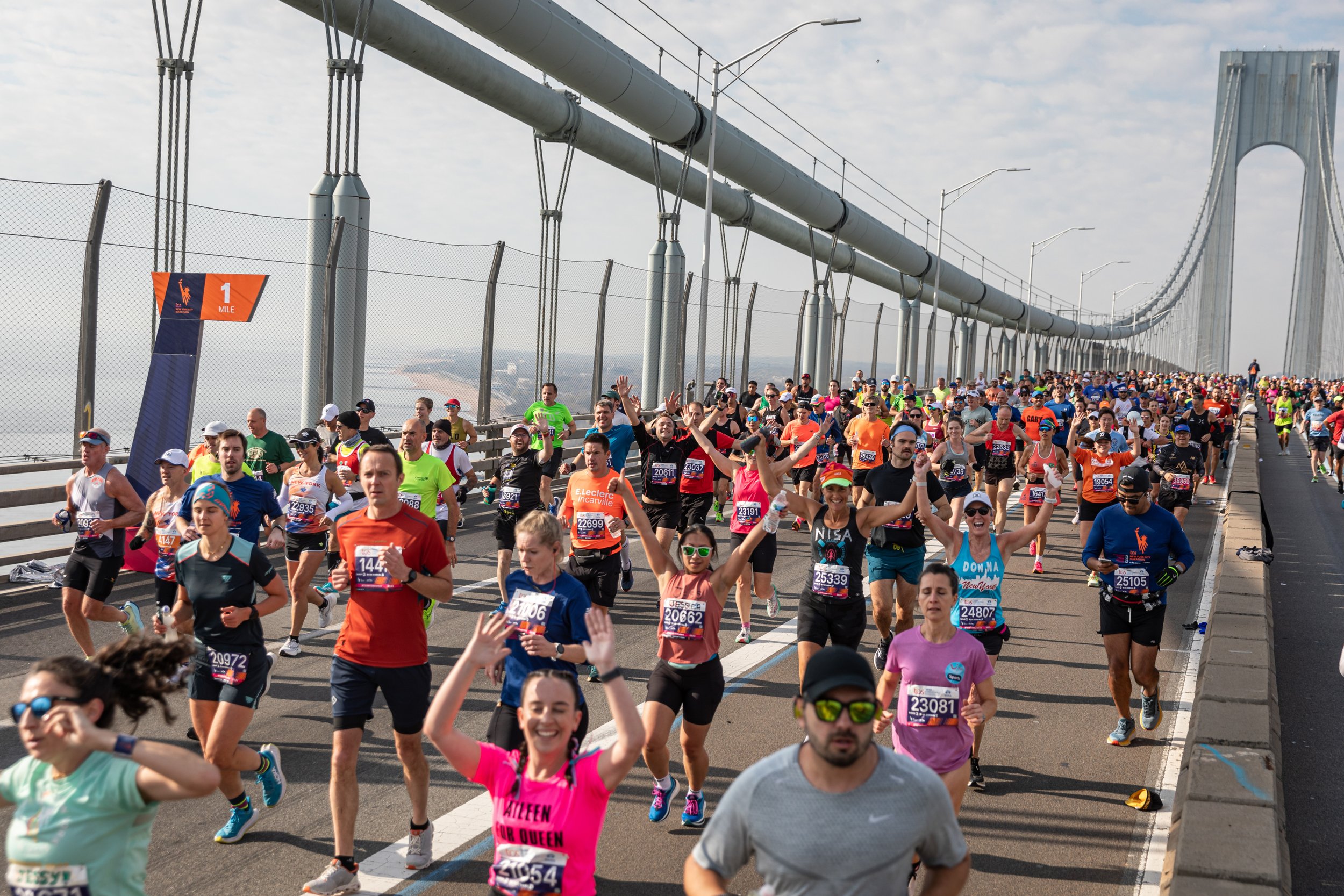 Numerous runners participating in a marathon or race, walking or jogging on a bridge with a large suspension tower in the background. Some runners are smiling and waving, wearing colorful athletic clothing and bib numbers.