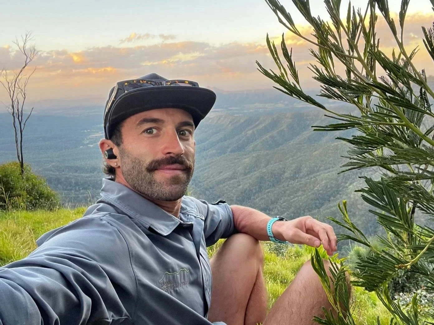 A man taking a selfie outdoors with a scenic mountain view at sunset in the background, dressed in outdoor gear, including a cap, jacket, and a fitness tracker, sitting on green grass near some trees and bushes.
