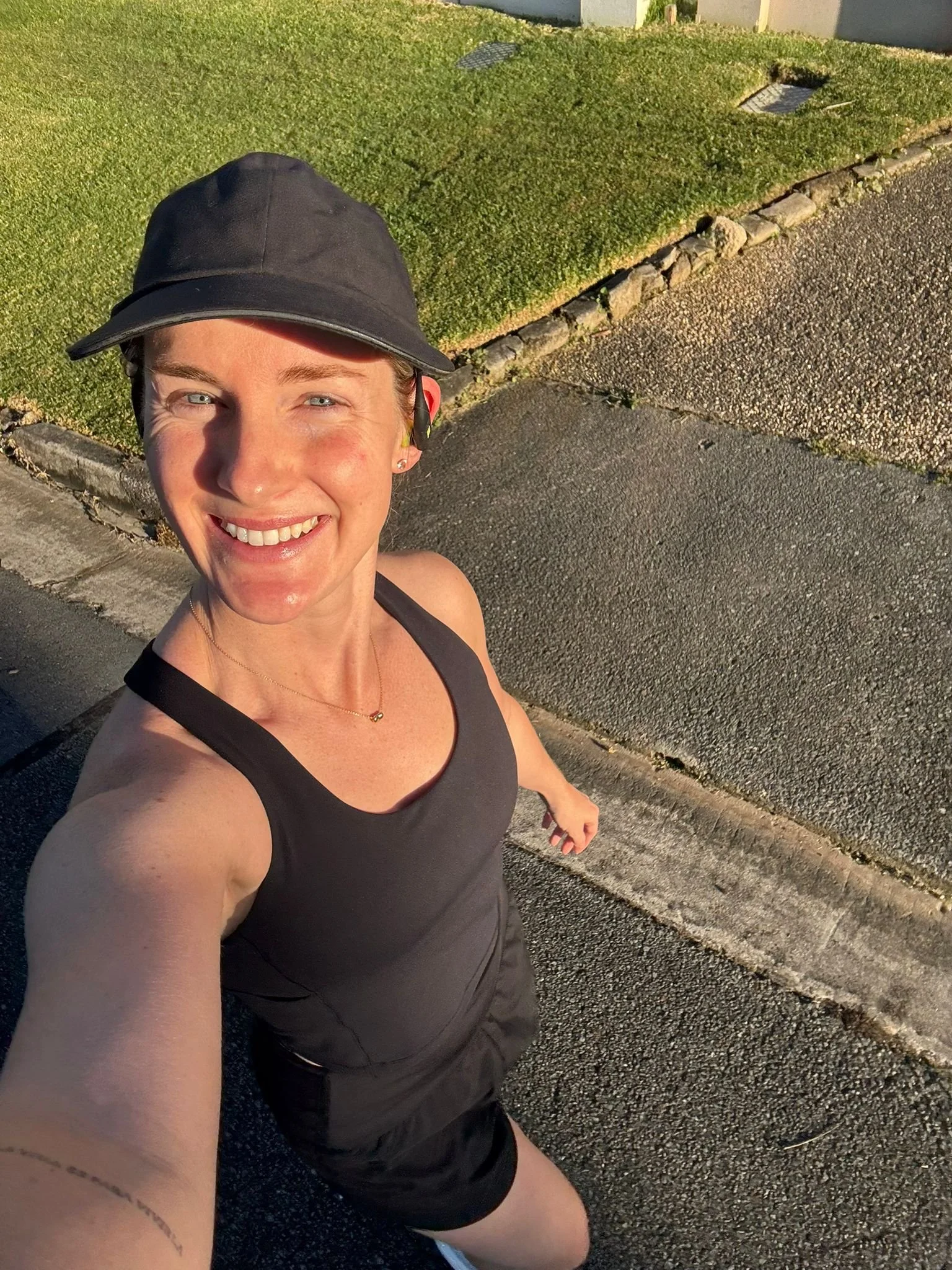 A smiling woman wearing a black cap and black athletic outfit taking a selfie on a sidewalk in the sunlight.