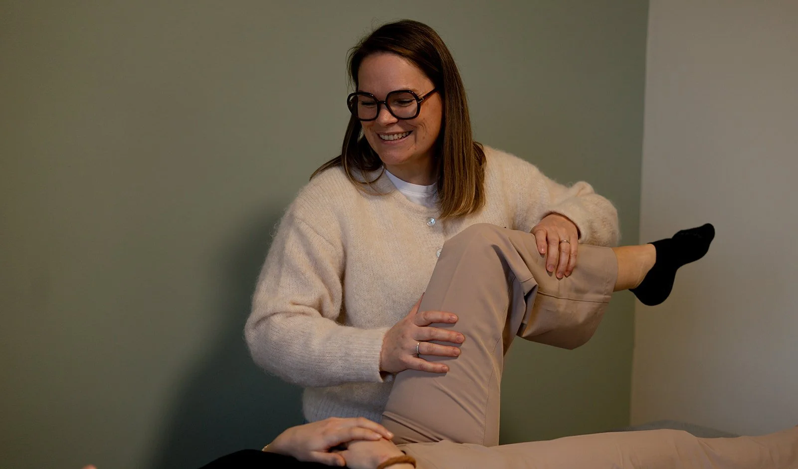 Une femme souriante aidant une autre personne à faire de l'exercice ou une thérapie physique, en soulevant sa jambe dans une pièce à lumière douce.
