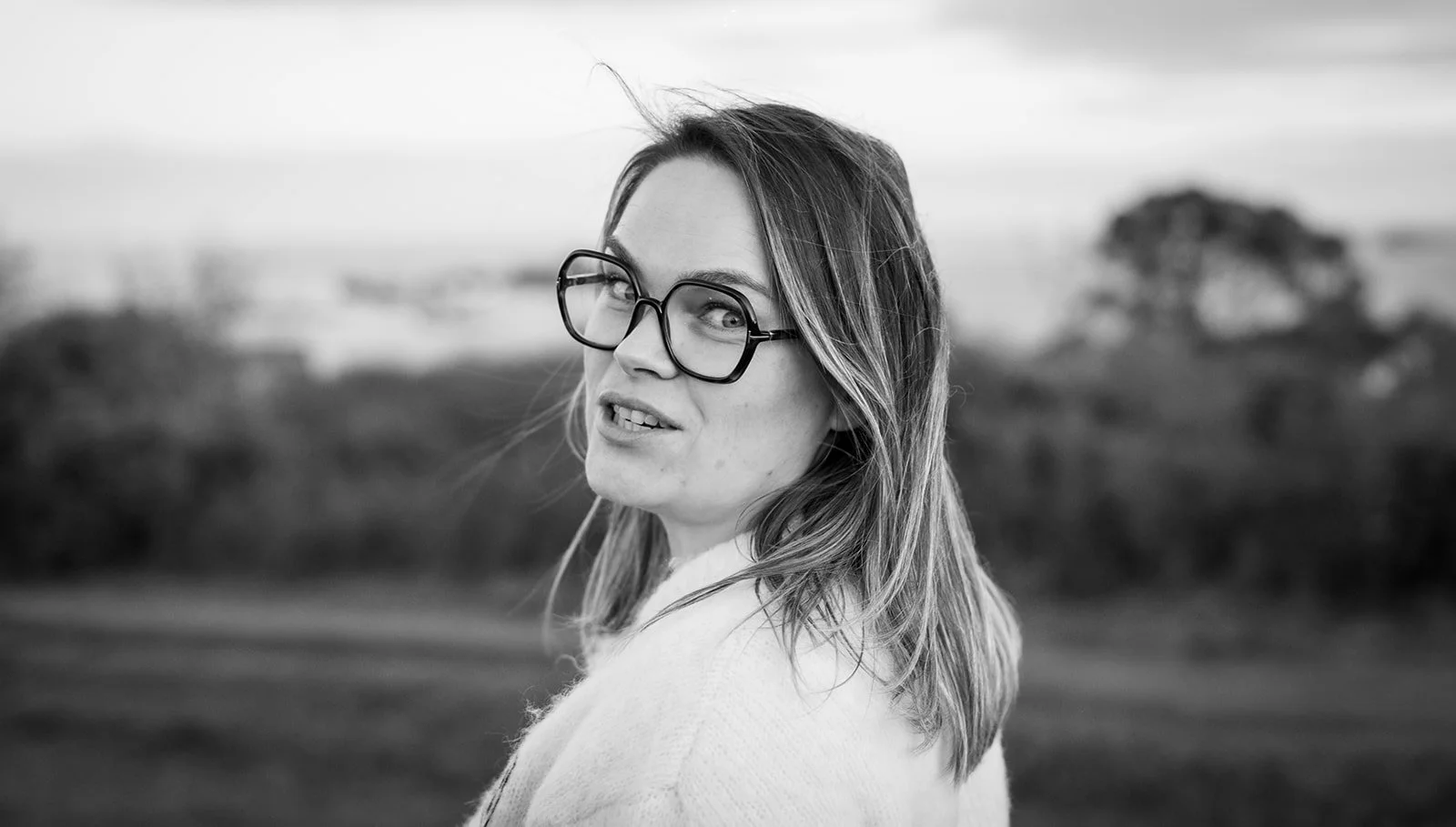 Une femme portant des lunettes regarde par-dessus son épaule dans un paysage naturel flou en arrière-plan, image en noir et blanc.