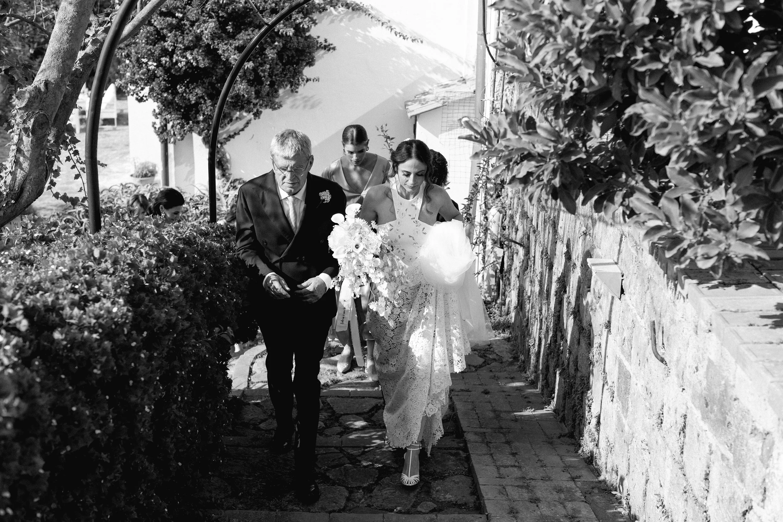 Un matrimonio cammina lungo un sentiero di pietra, circondato da piante e fiori, sotto la luce del sole. La sposa indossa un abito di pizzo chiaro e tiene un bouquet, mentre il uomo accanto a lei è in abito scuro.