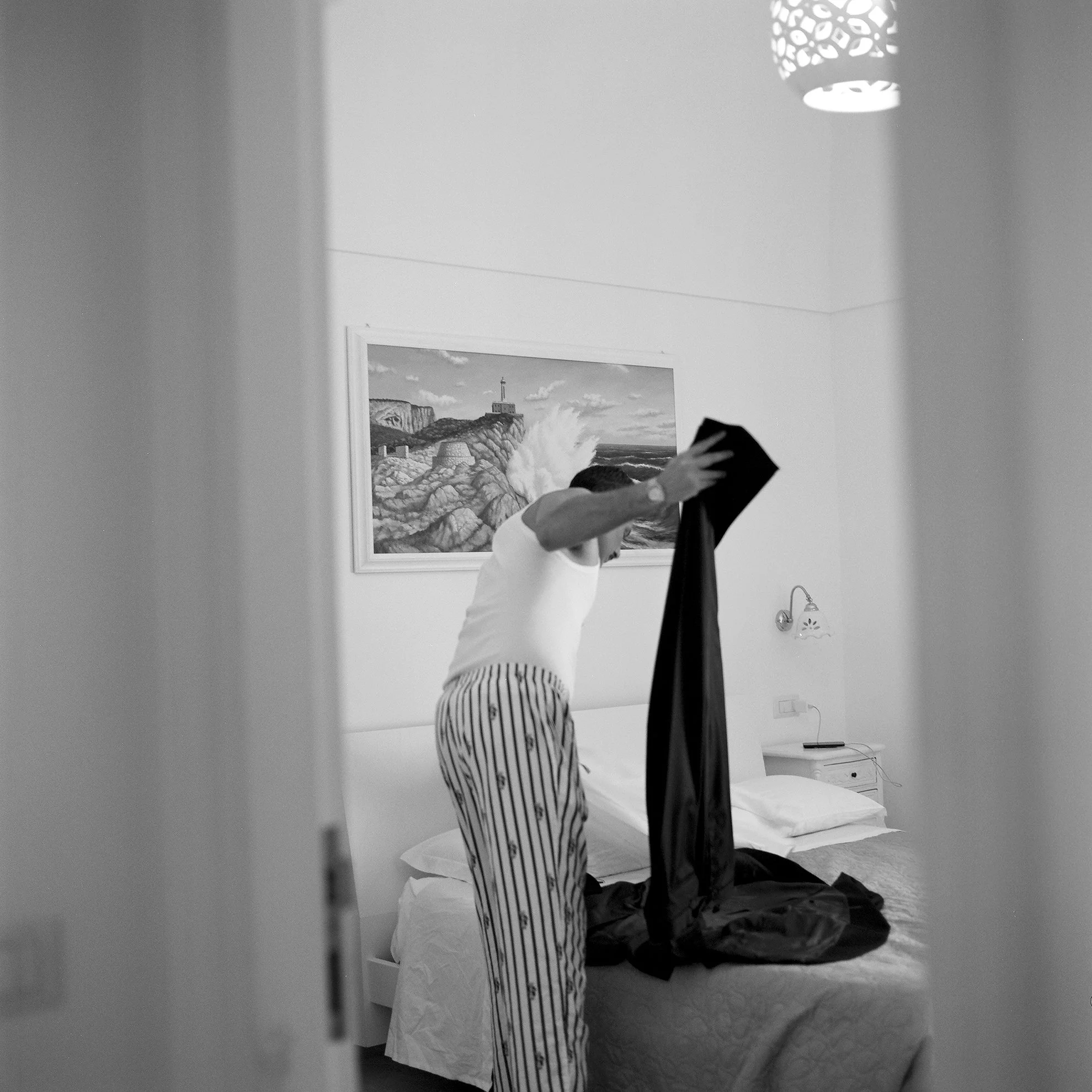 Una persona che si prepara a vestirsi in una camera da letto, vista attraverso una porta aperta, con una parete decorata con un quadro e un lampadario