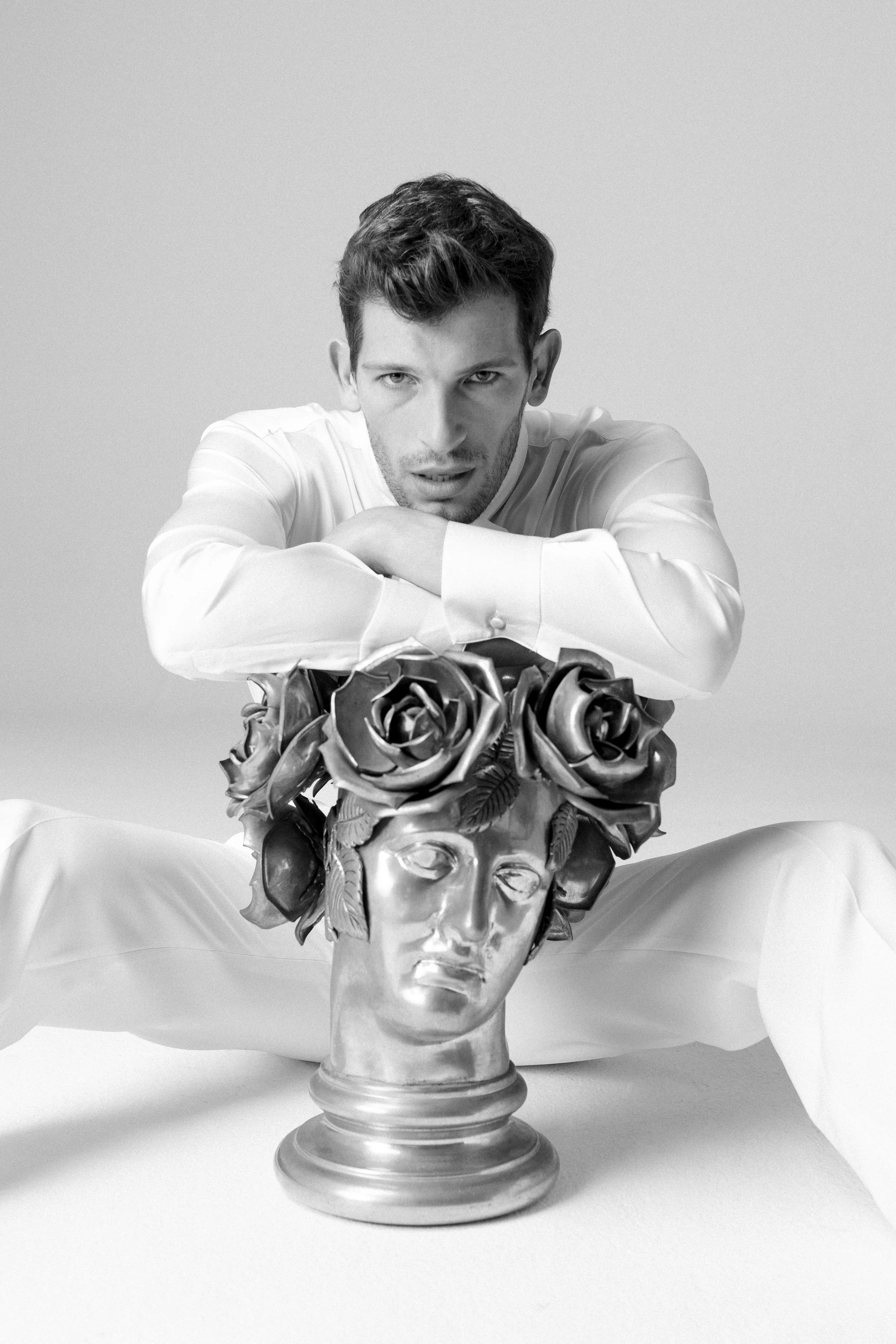 Un uomo con camicia bianca seduto davanti a una statua di una testa umana decorata con fiori e foglie, in stile artistico.