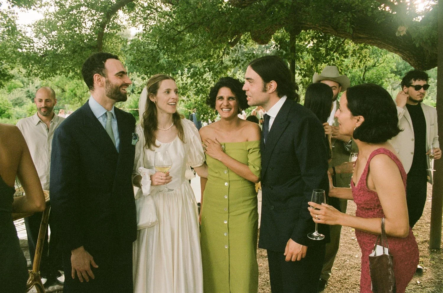 Gruppo di persone che socializzano in un matrimonio all'aperto, sorridenti e che tengono bicchieri di vino, sotto un albero verde.