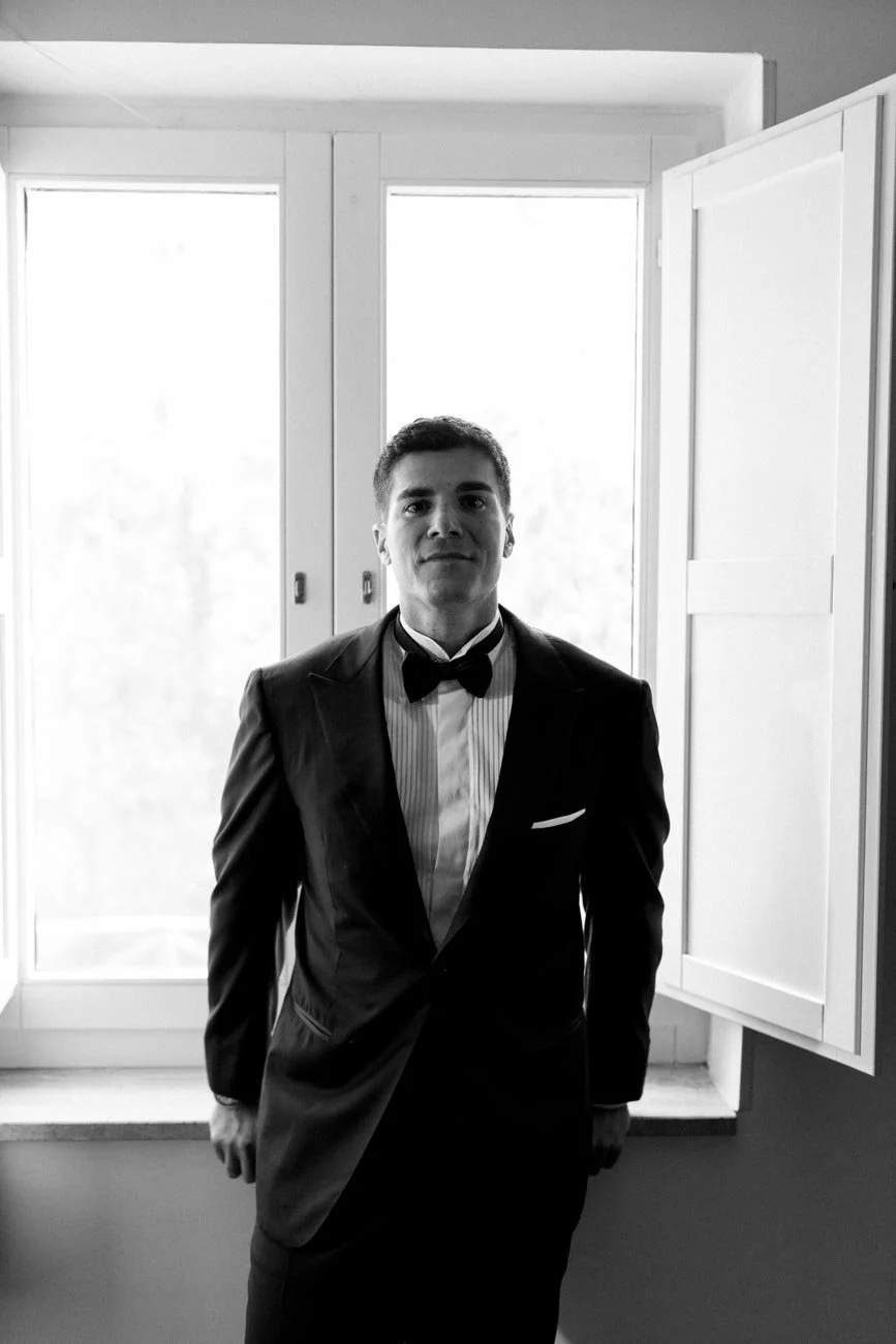 Un uomo vestito in modo formale con abito nero e papillon si trova davanti a una finestra luminosa, in una stanza con mobili bianchi.