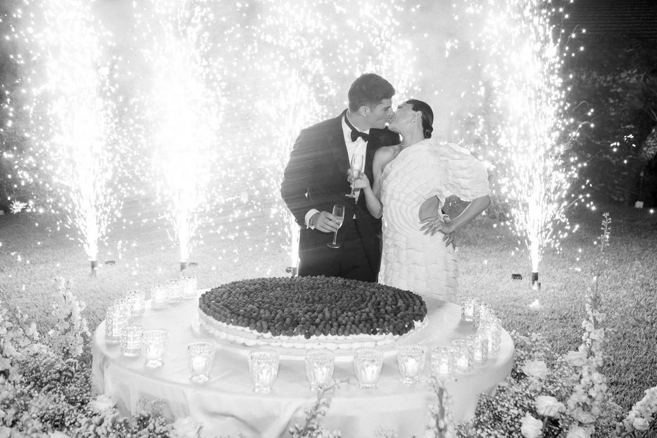 Una coppia di sposi si bacia davanti a una torta nuziale e fuochi d'artificio in un giardino.