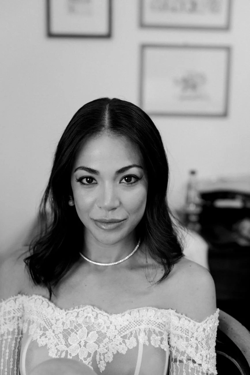 Ritratto di una donna con abito di pizzo, capelli scuri portati sciolti, indossa una collana di perle, sfondo interno con quadri alle pareti.