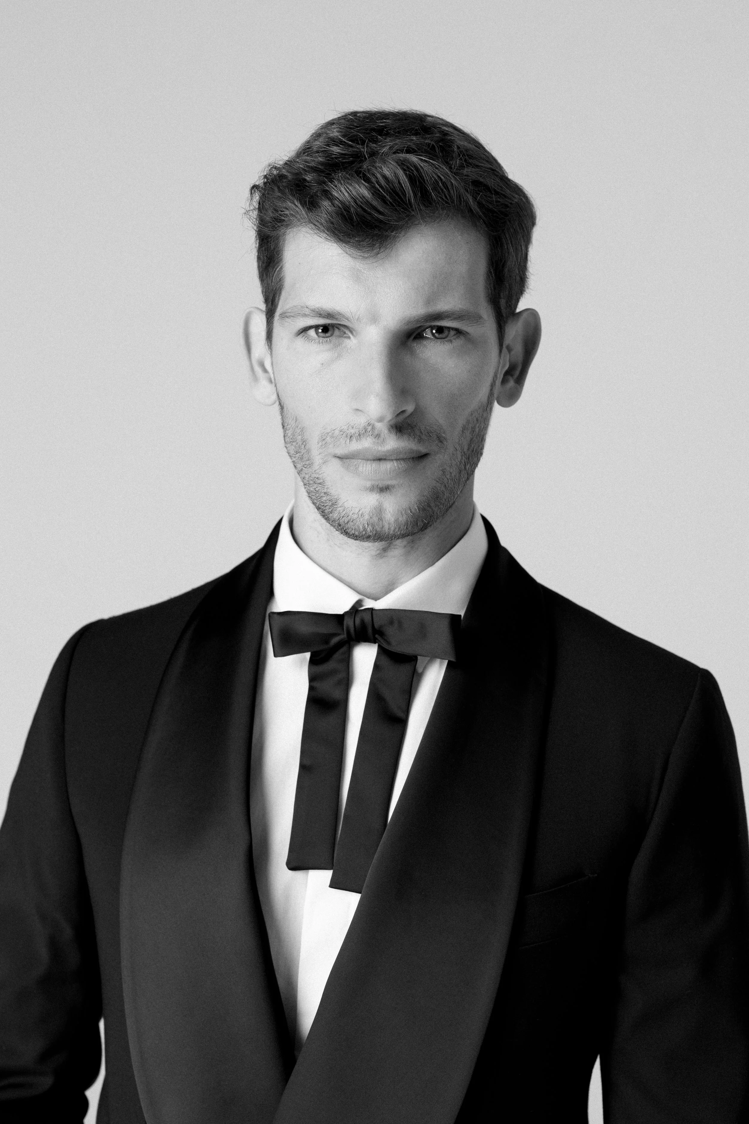 Ritratto di un uomo elegante con giacca nera, camicia bianca e fiocco nero, che guarda verso la fotocamera, in un ambiente semplice.