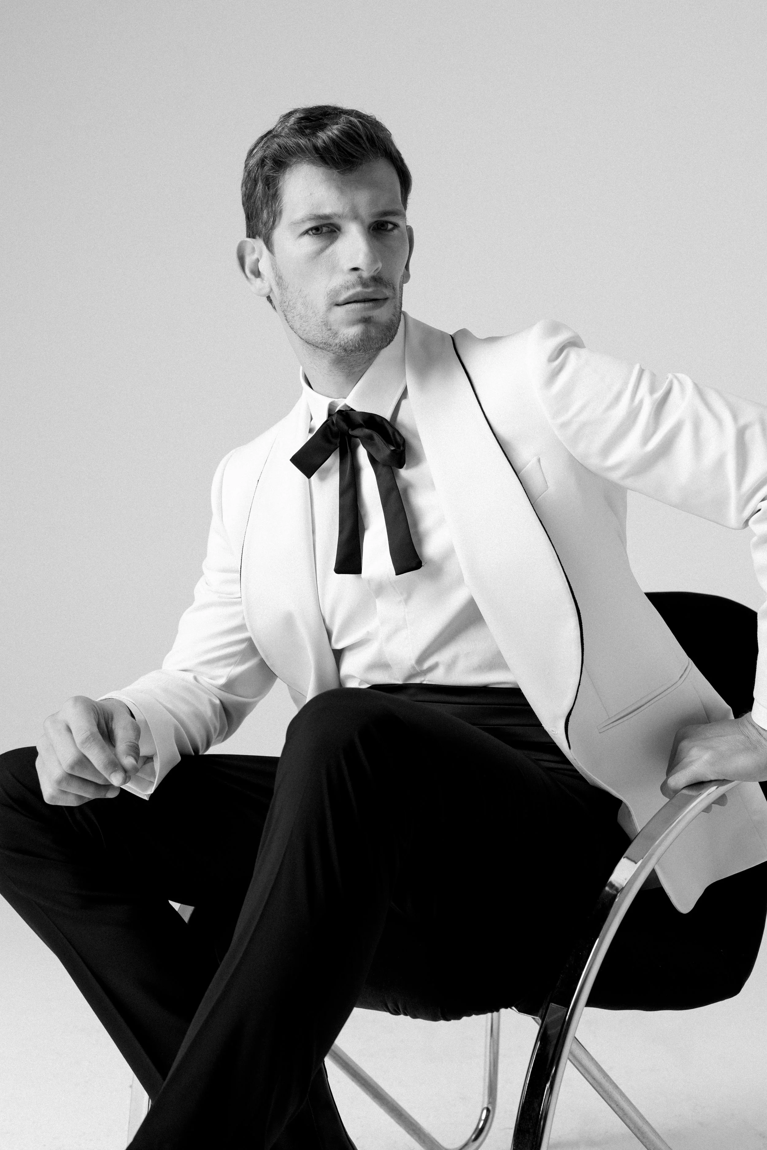 Uomini seduto su una sedia, indossa un abito bianco con dettagli neri, camicia bianca e una cravatta nera a fiocco, in un ambiente neutro.