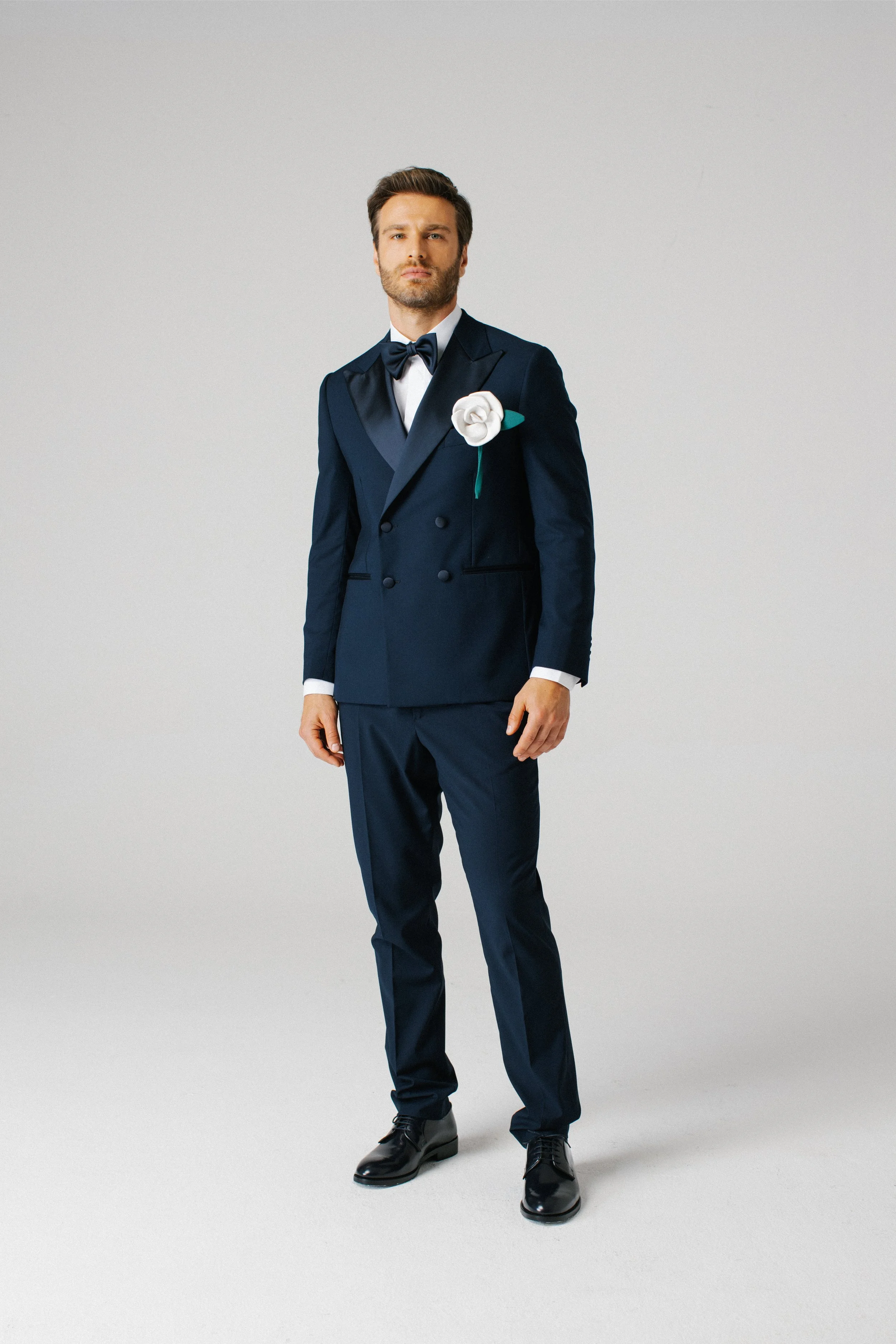 Uomo vestito in abito da sera blu con papillon e boutonnière bianco, in piedi contro uno sfondo grigio chiaro.