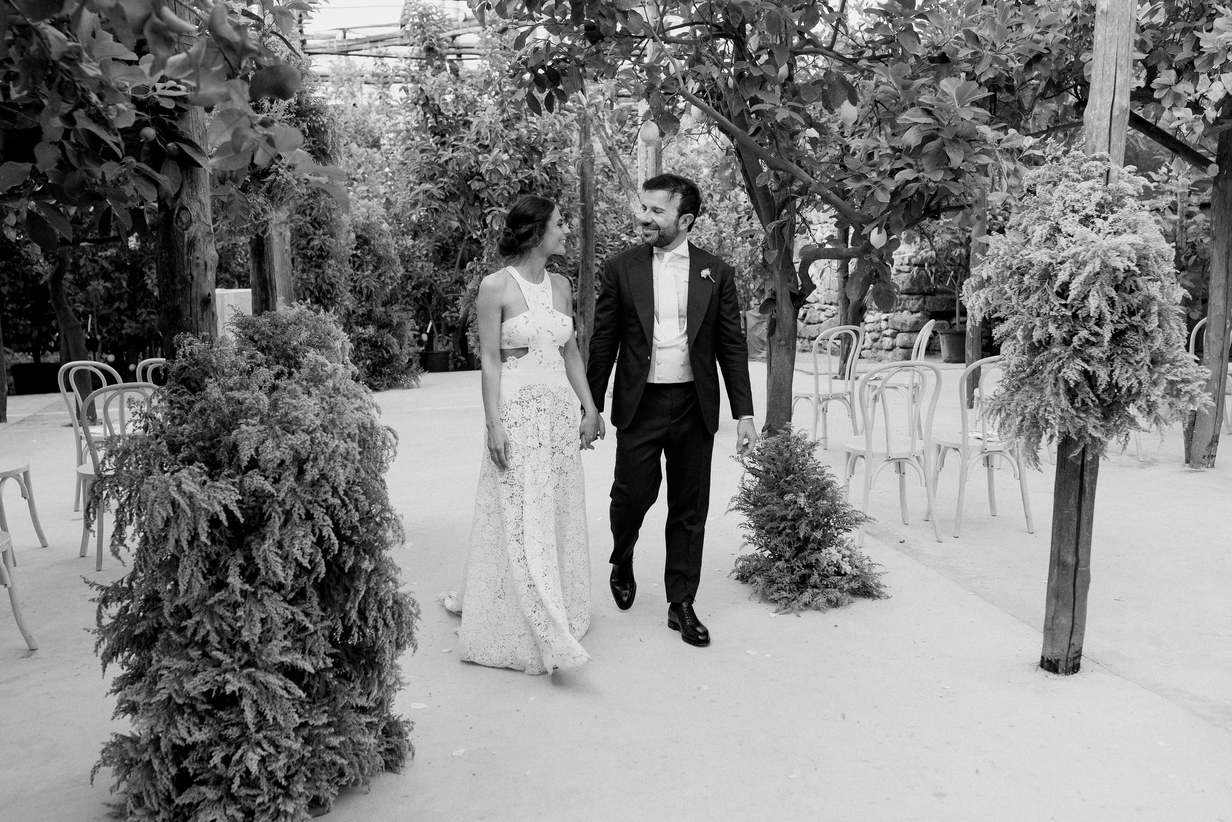 Una coppia di sposi si tiene per mano e si guarda negli occhi in un luogo adornato con piante e sedie vuote.