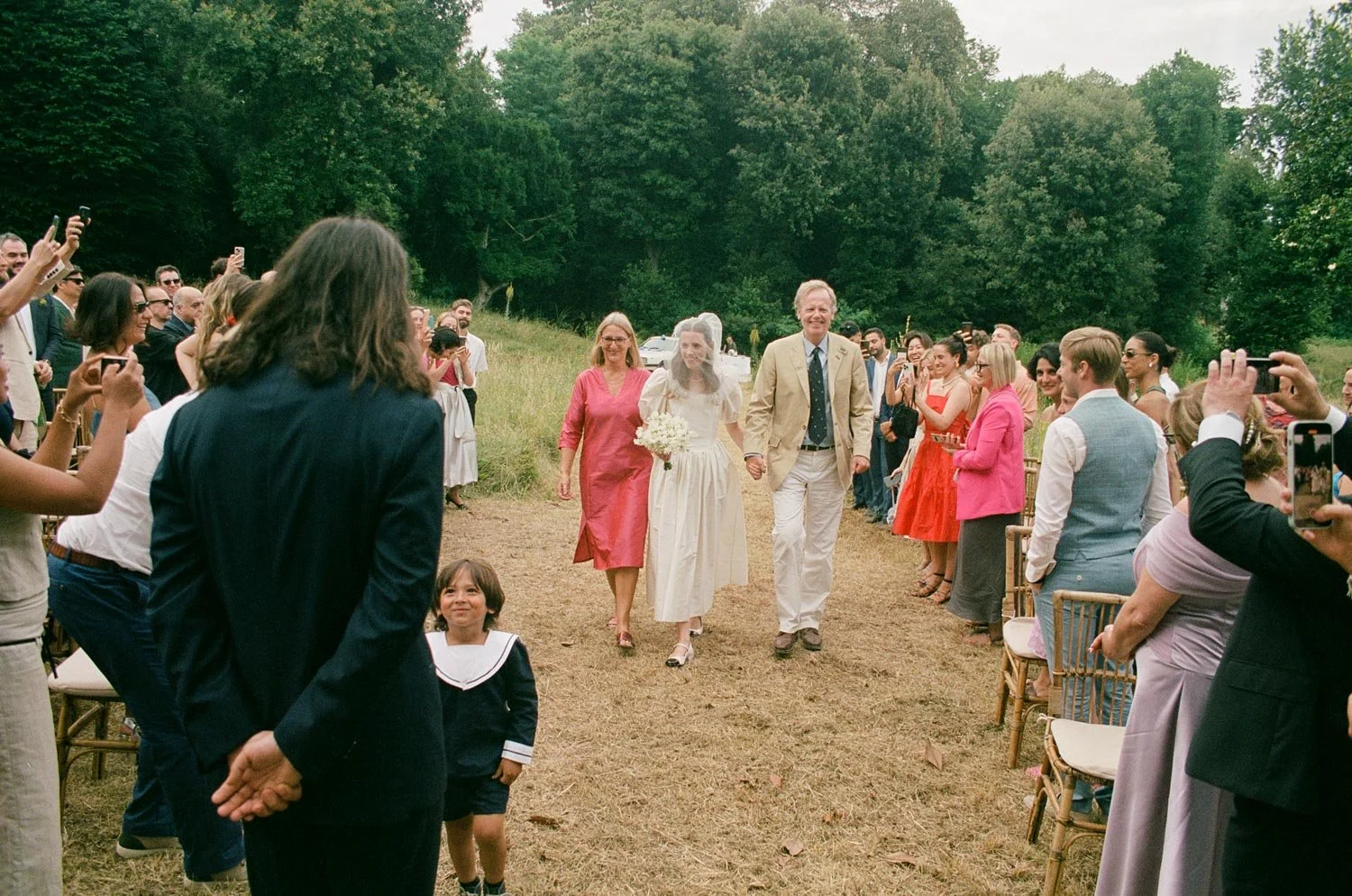 Una coppia di sposi entra in un evento all'aperto, circondati da amici e parenti che li applaudono e scattano foto. L'atmosfera è gioiosa e celebrativa, con molte persone sorridenti e relative decorazioni.
