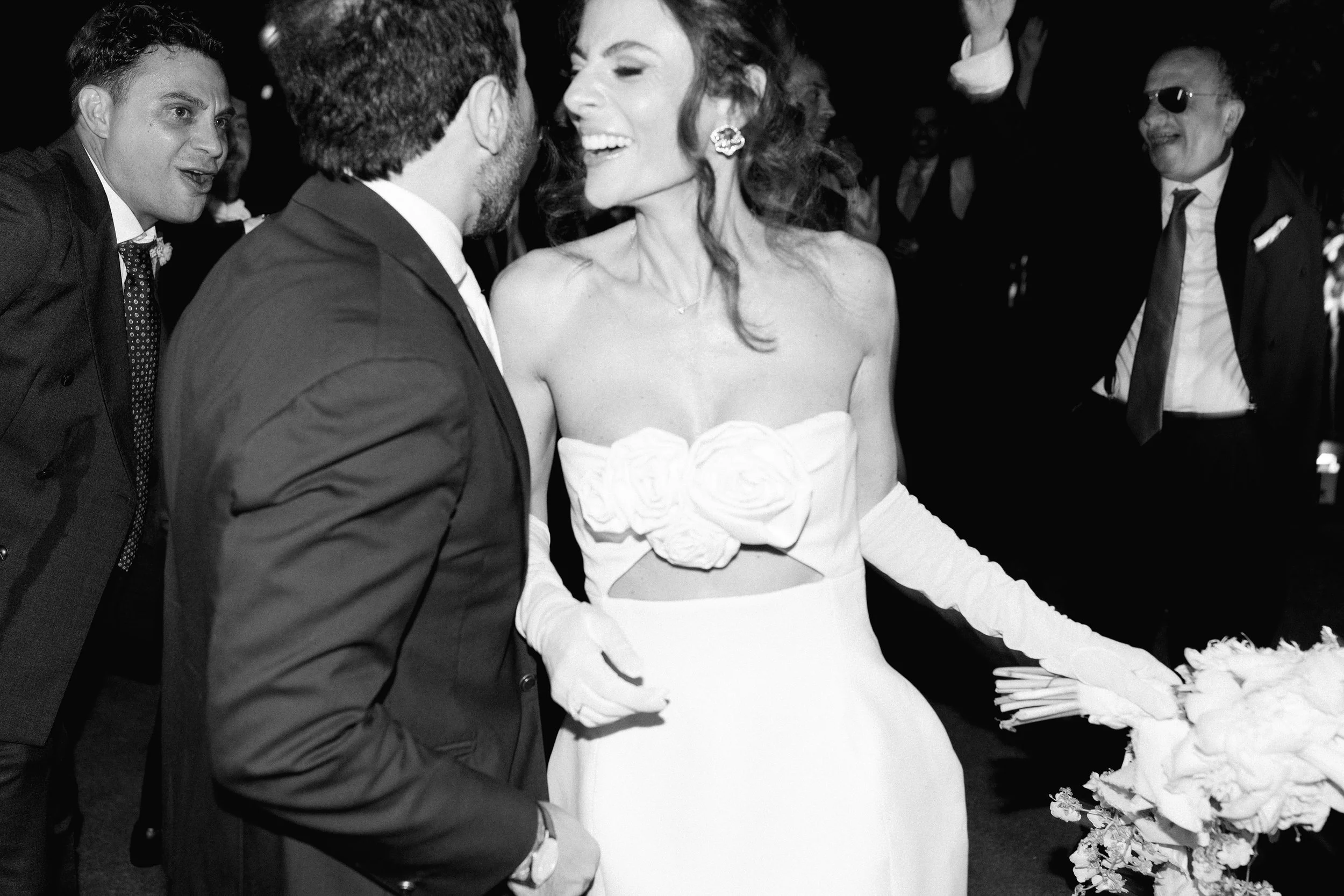 Una coppia di sposi si bacia sorridendo, circondati da amici in festa, in un evento elegante.