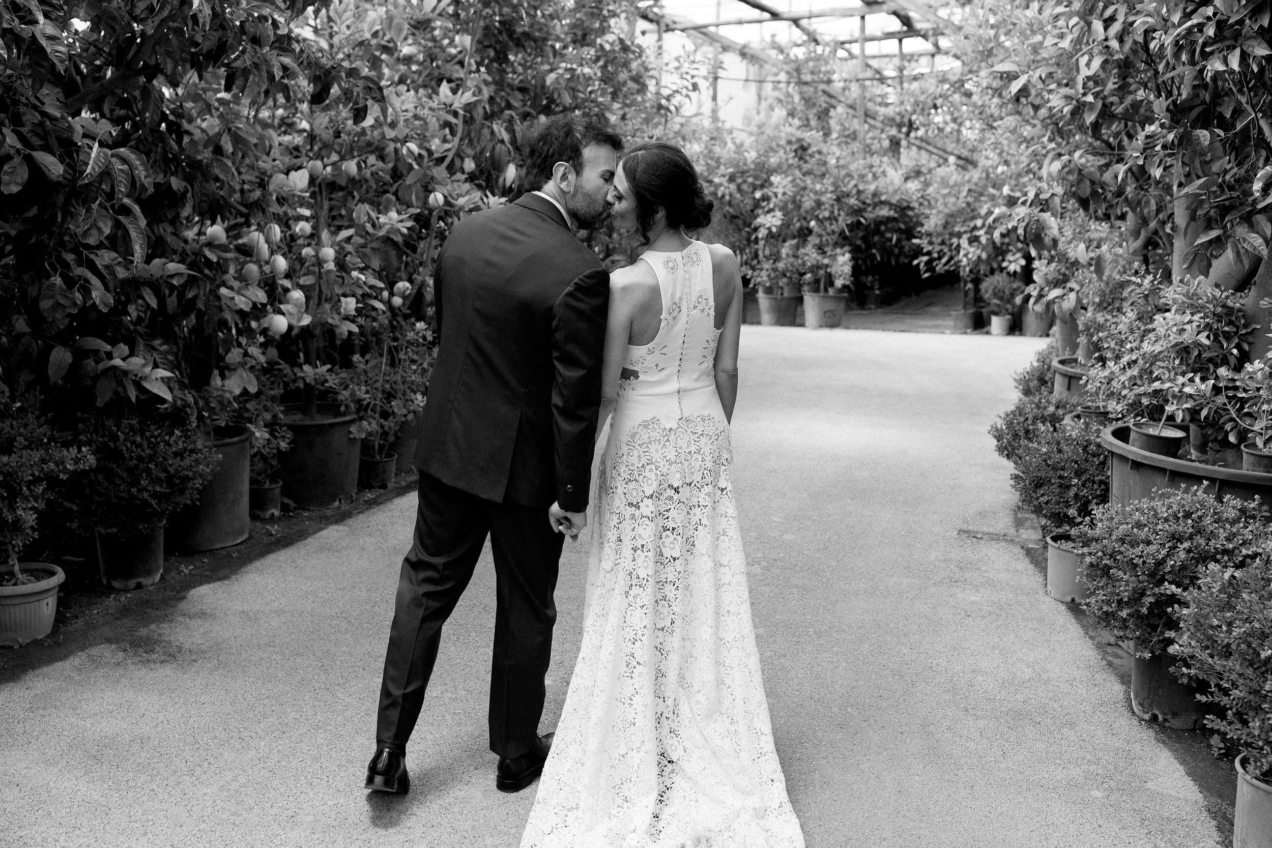 Una coppia in abiti da sposa e sposo si bacia con il naso e la fronte che si toccano, camminando in un giardino.